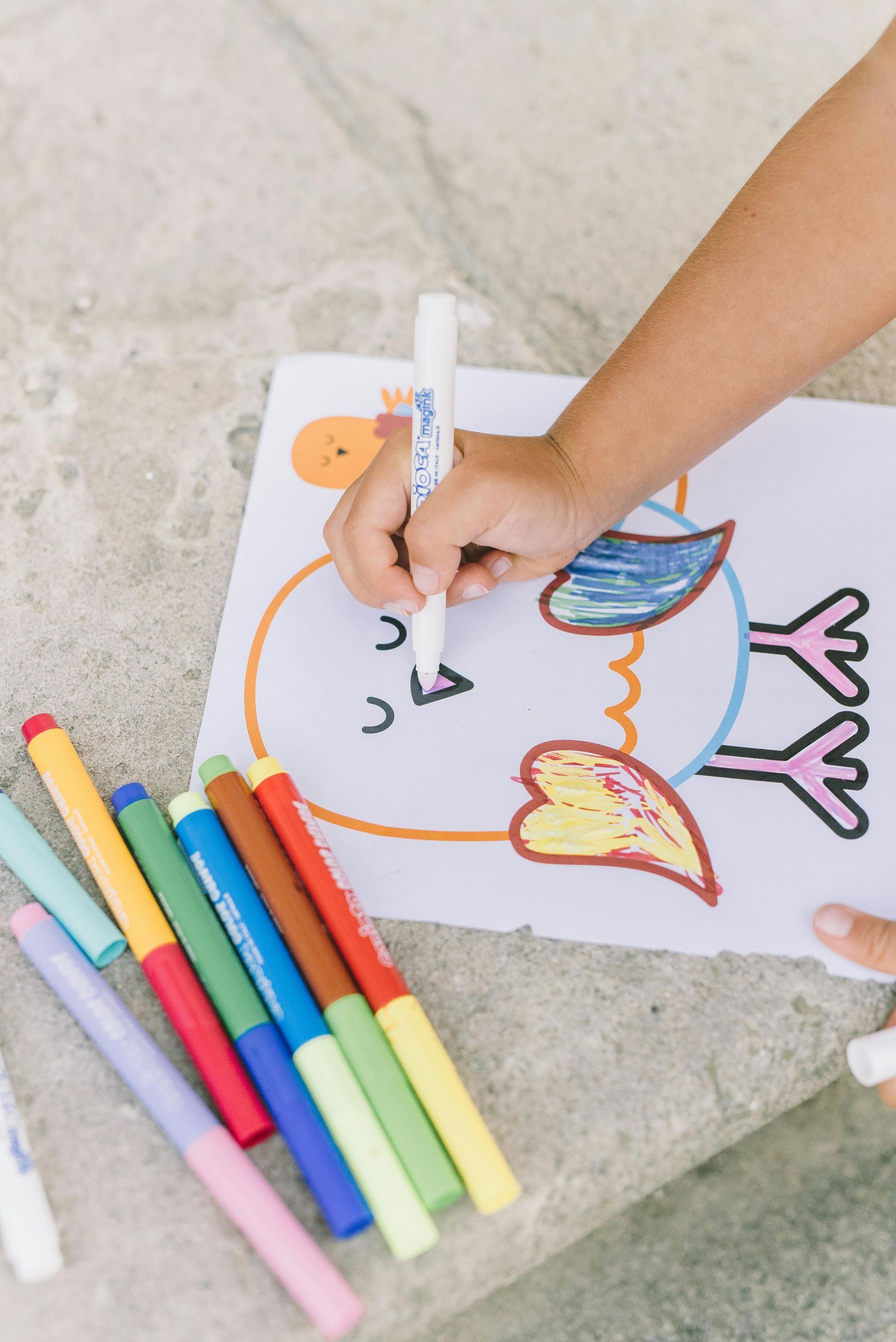 Child's hand coloring a bird drawing with a white marker. Colorful markers are next to the artwork.
