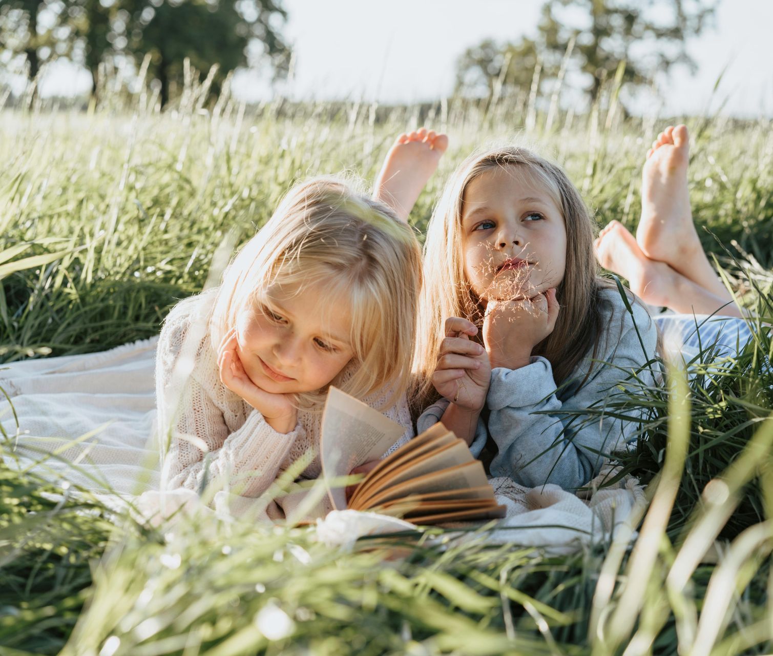 Two young girls lie in tall grass, reading a book.