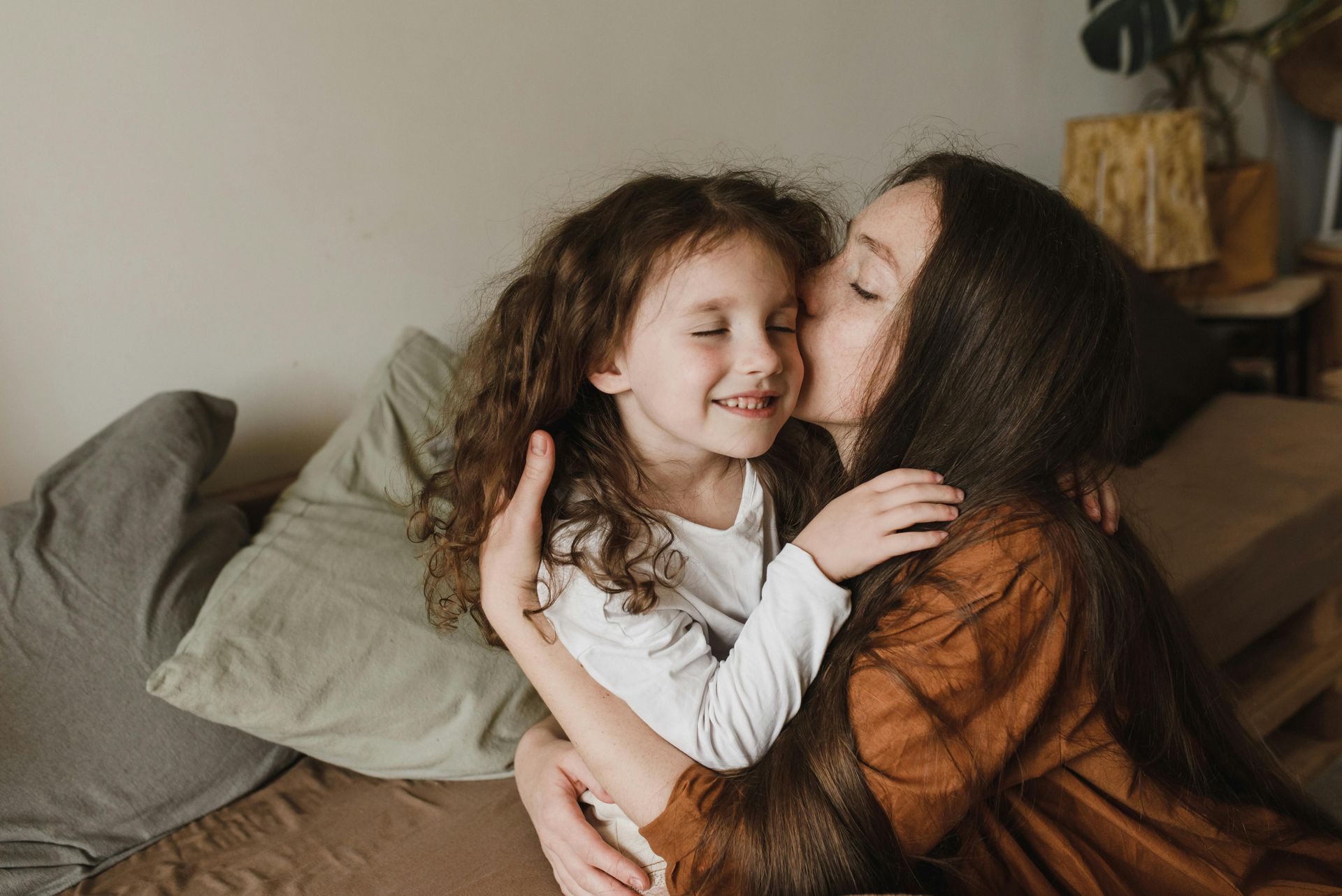 A girl kisses another girl on the cheek. They are indoors on a bed, smiling.