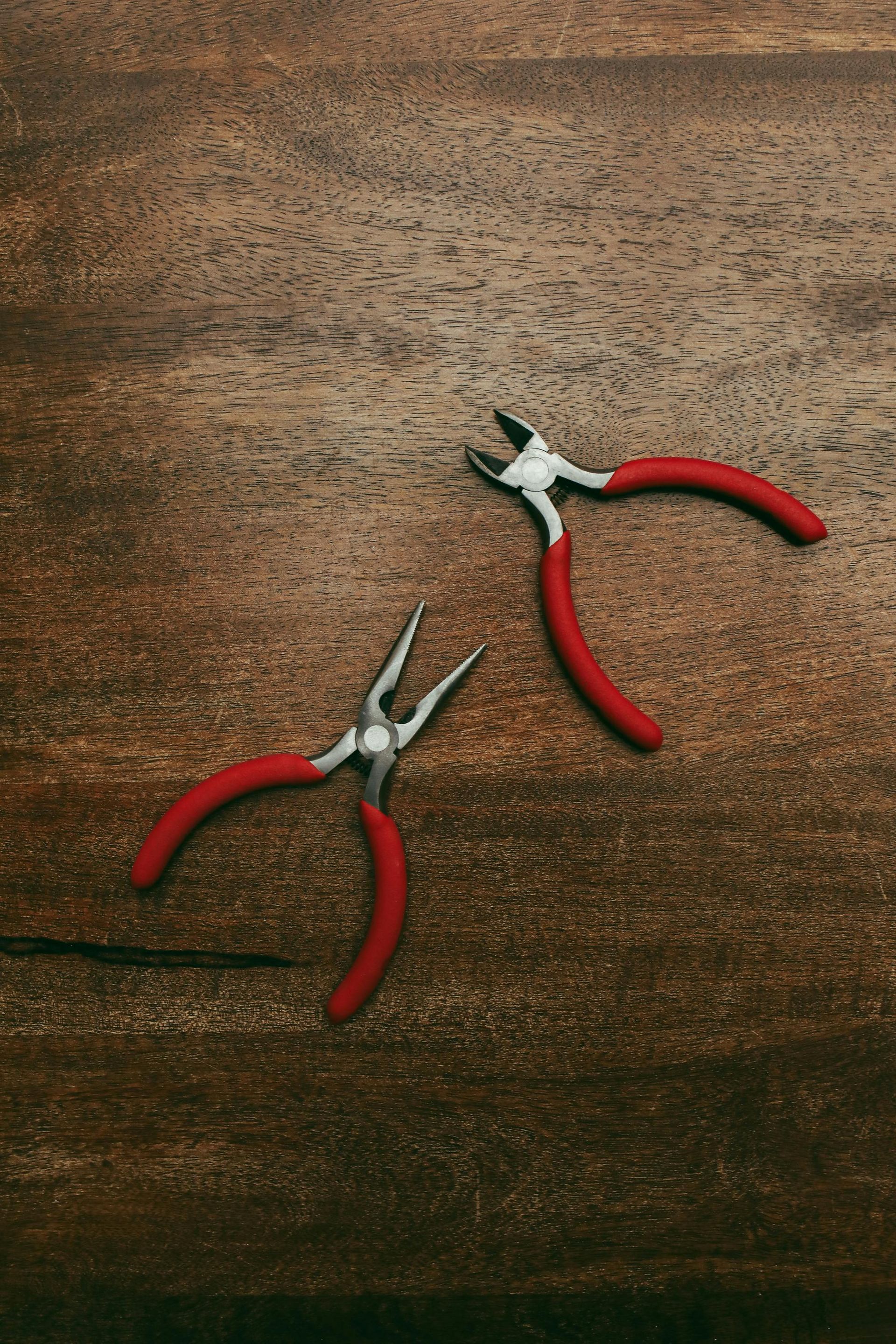 Two red-handled pliers on a dark brown wooden surface.