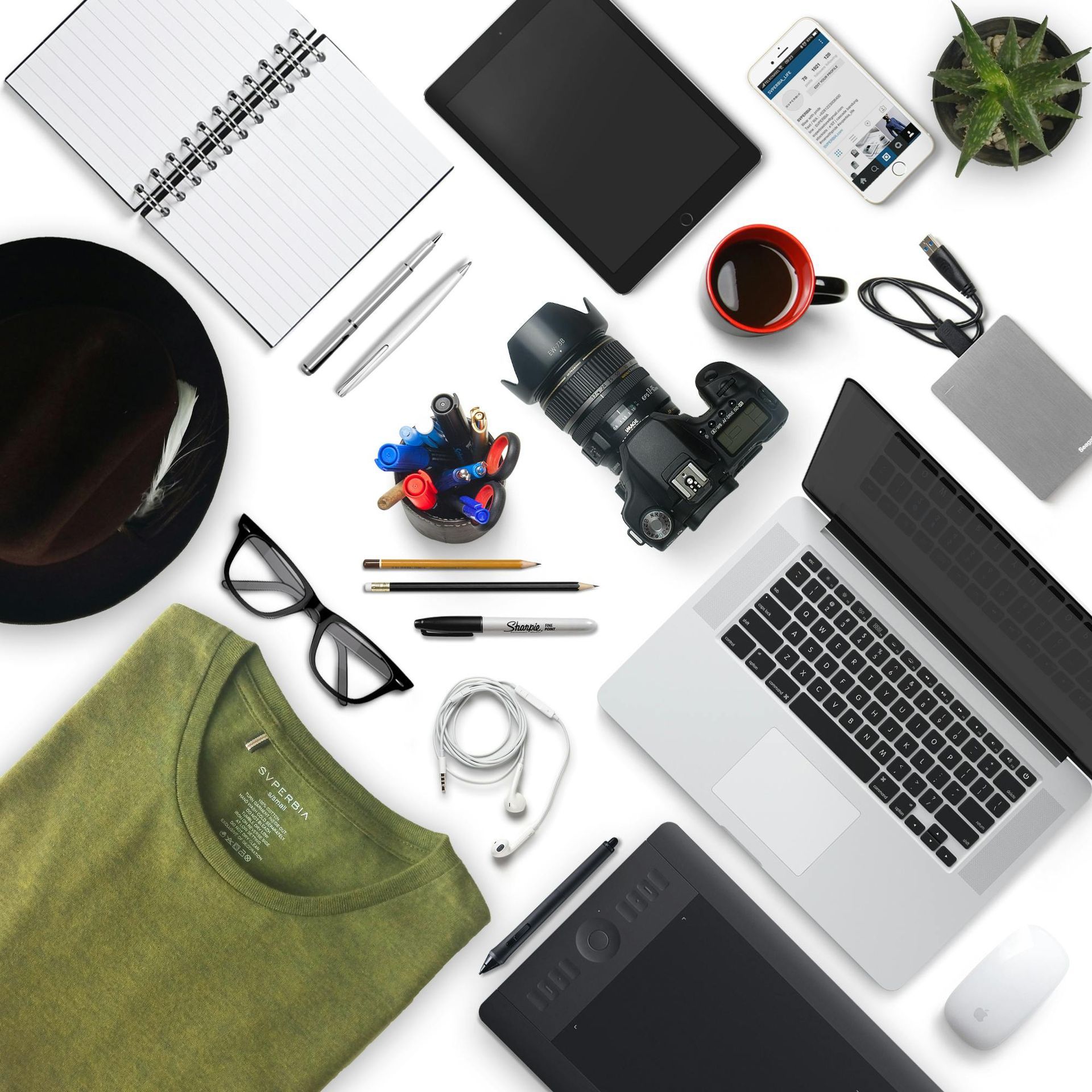 Overhead shot of desk with laptop, camera, tablet, notebook, shirt, and other office supplies.