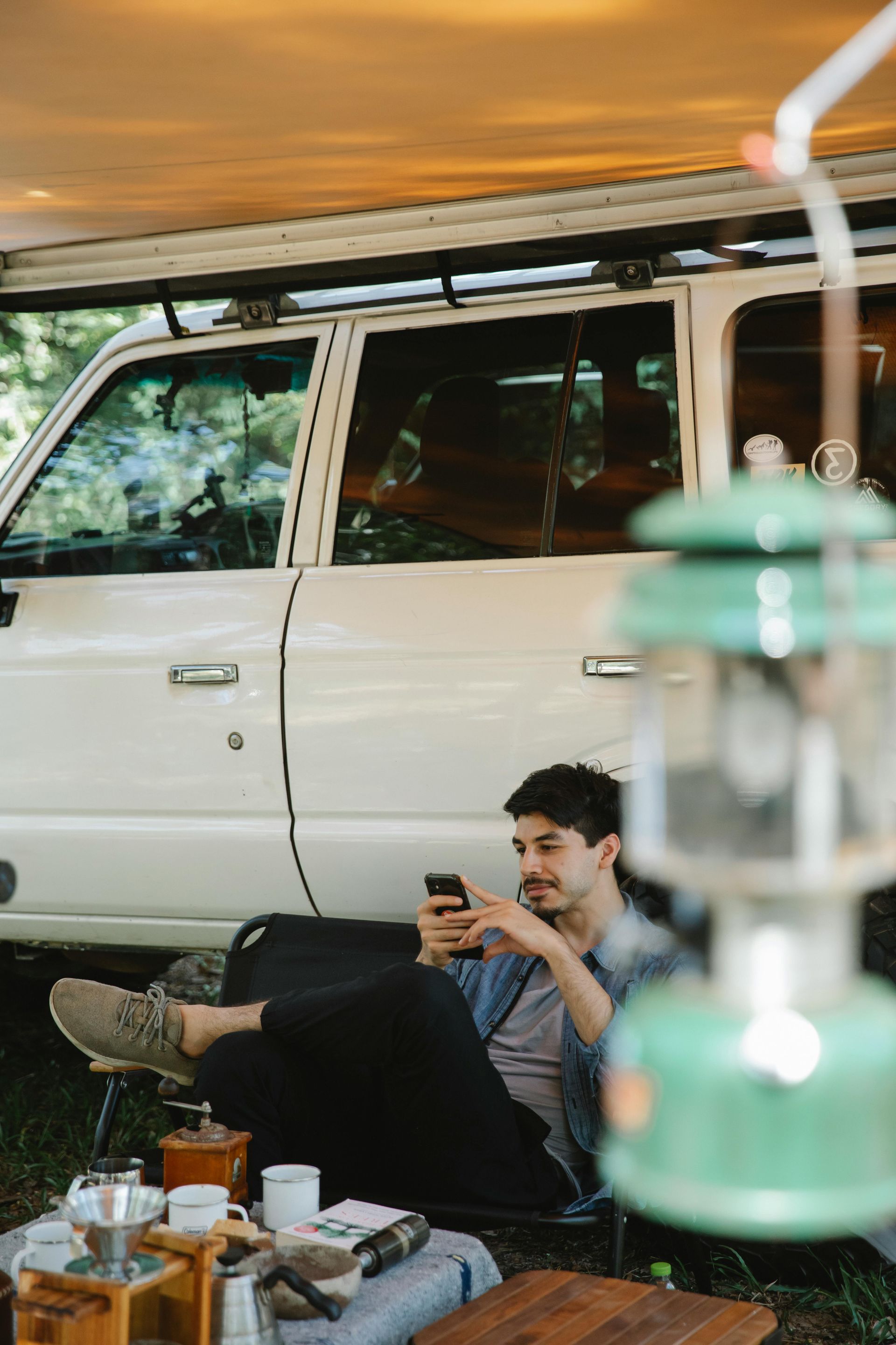 Man uses phone, relaxed by a white SUV with awning. Camping gear and green lantern in foreground.