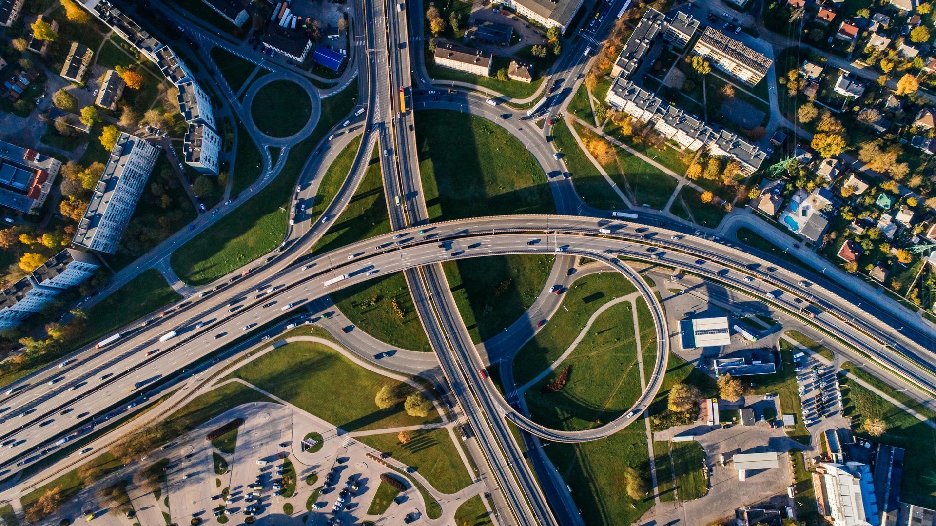 Overhead view of a multi-level highway interchange with many cars, green space, and surrounding buildings.