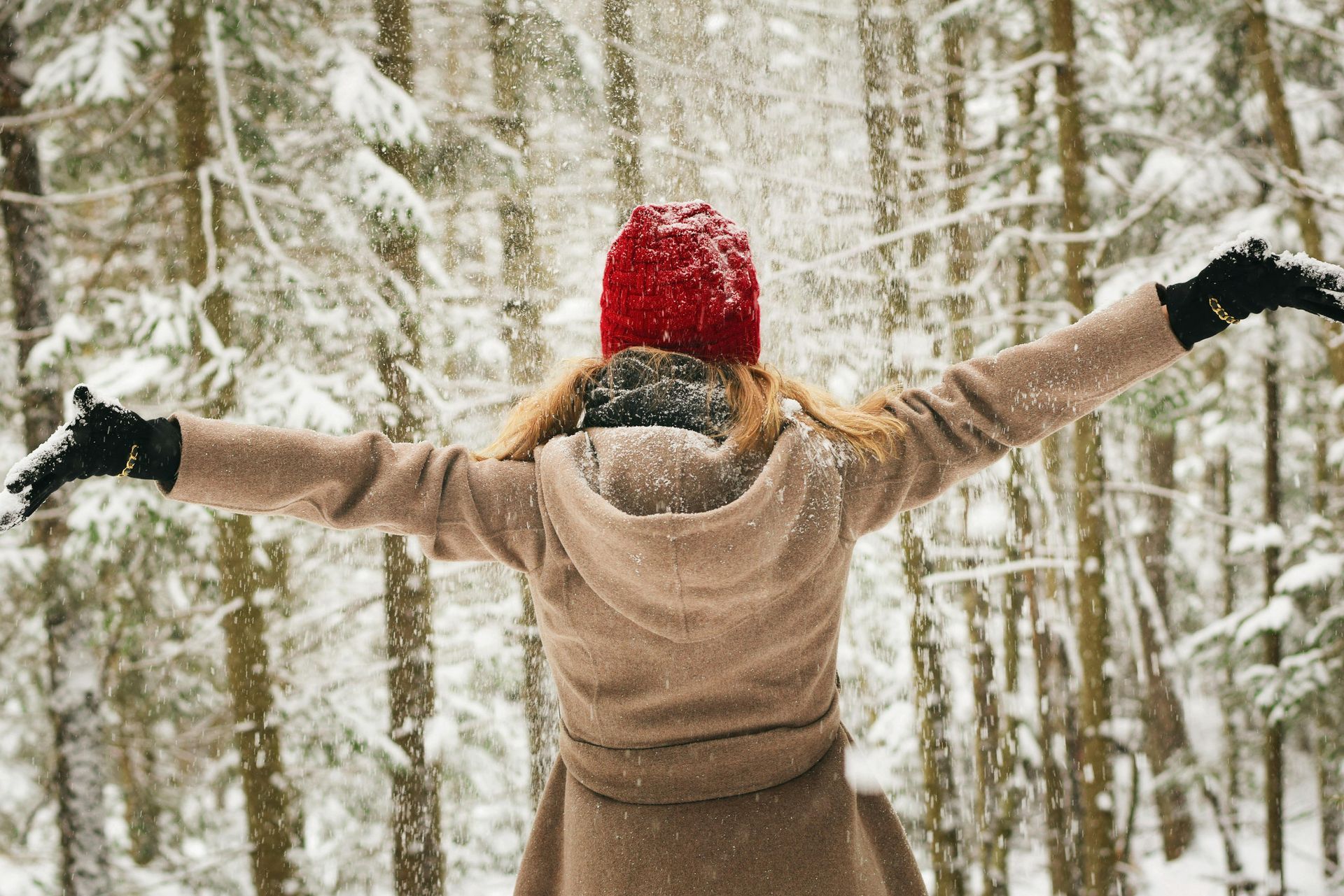 Woman in red hat and beige coat with arms outstretched in a snowy forest.