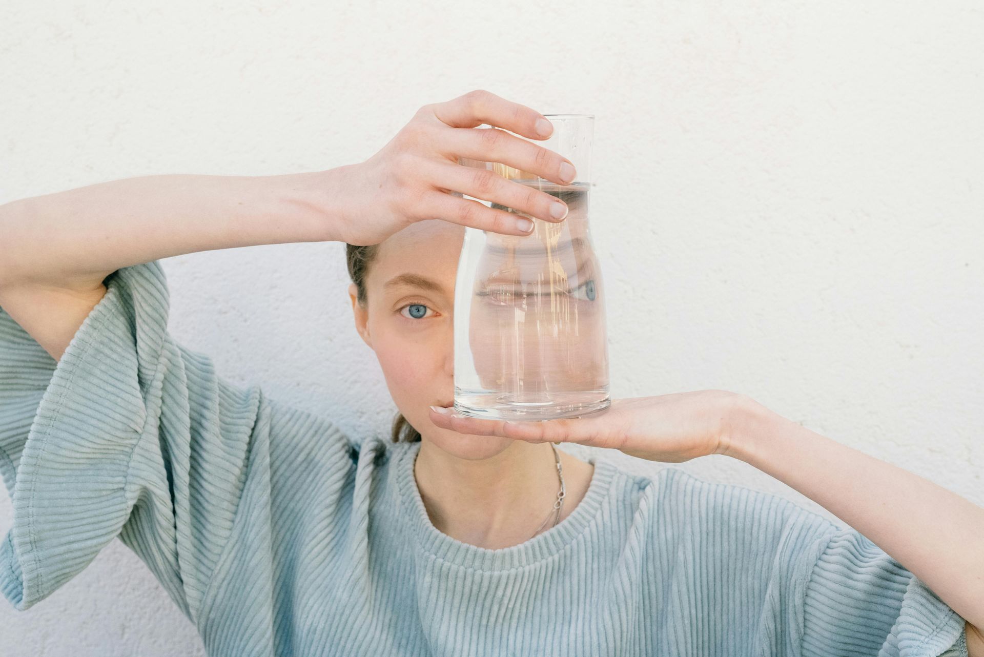 Woman holding a tall glass of water in front of her face, against a white wall.