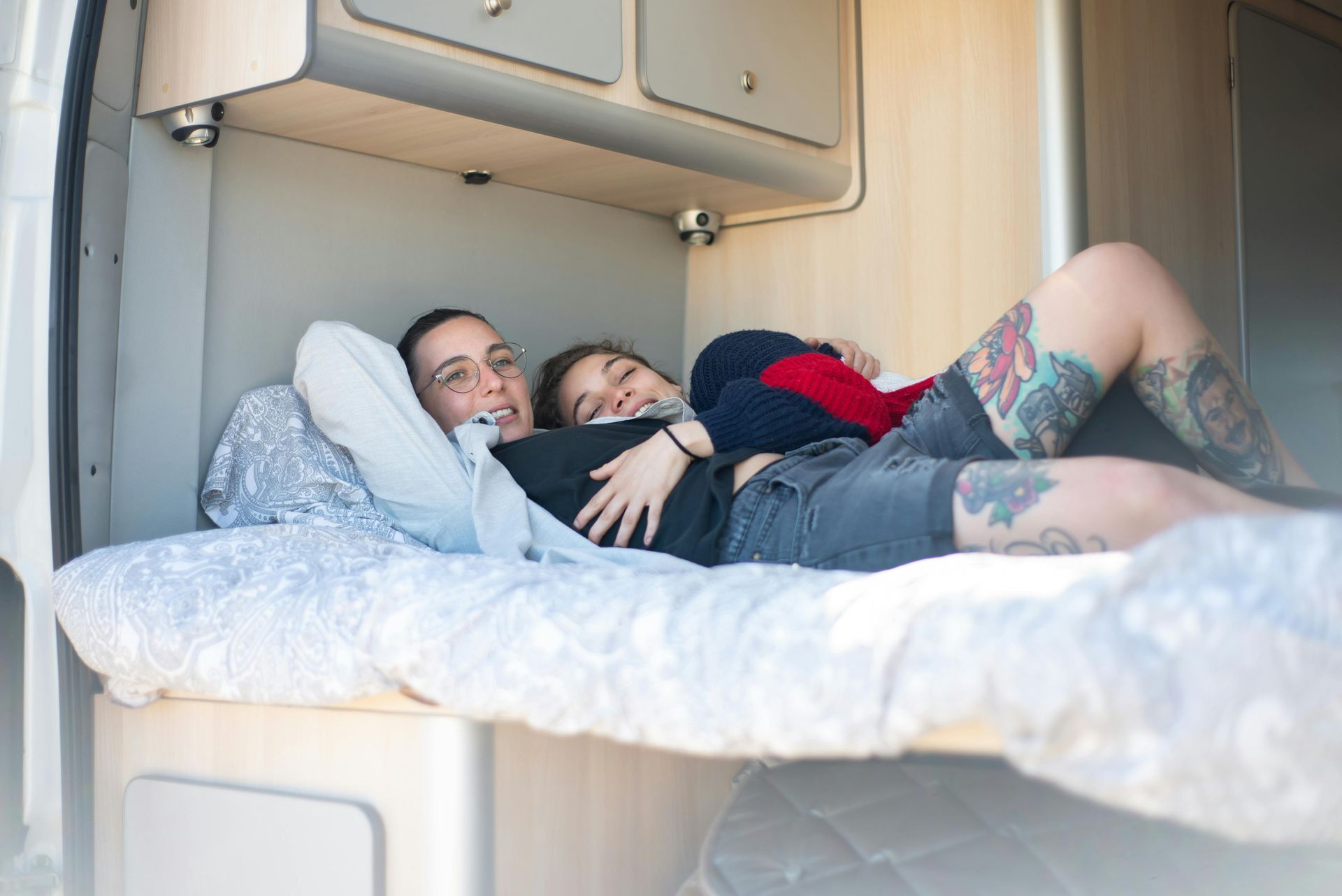 Two people lying on a bed in a camper van, smiling. One has arm tattoos. Beige cabinets above.