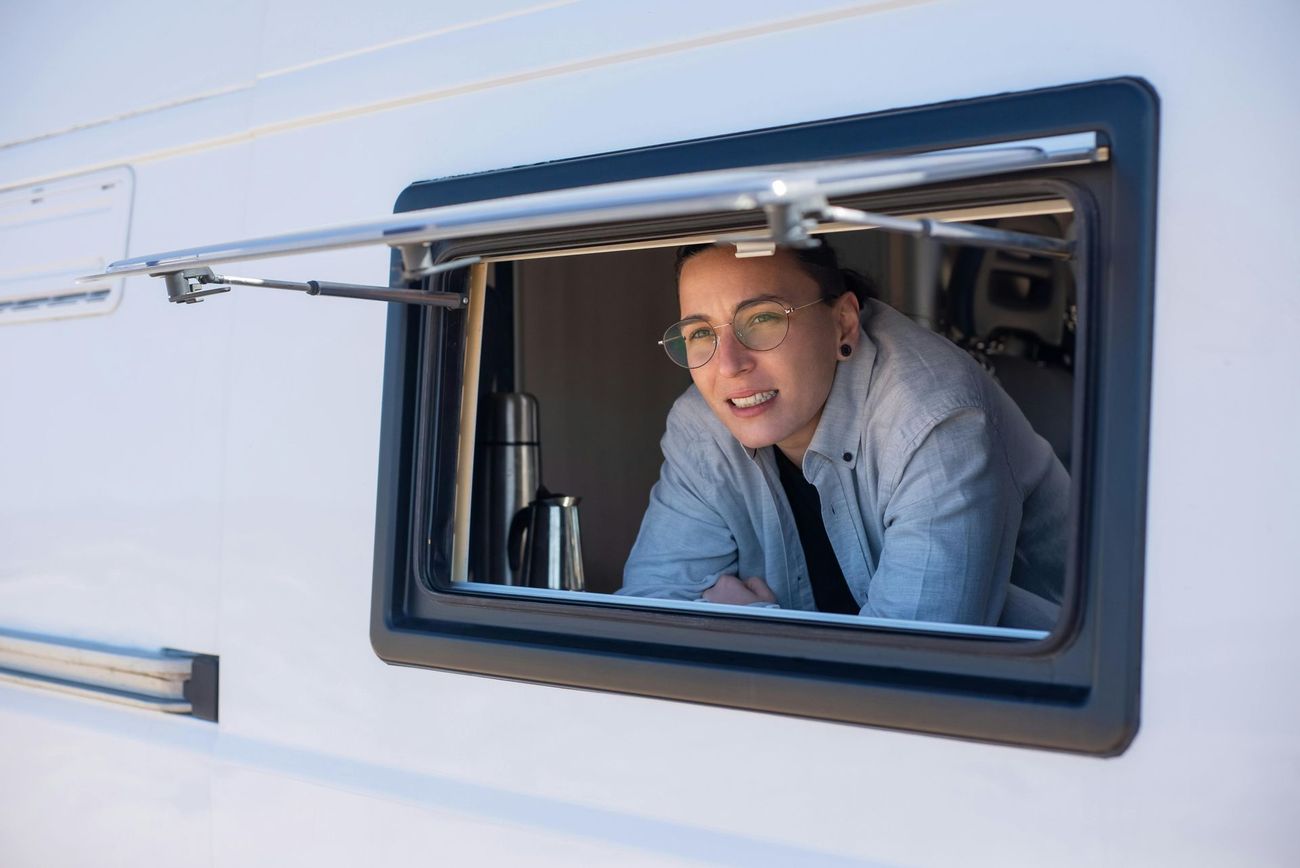 Person in glasses leaning out of a camper window, looking to the side. Silver thermos visible.