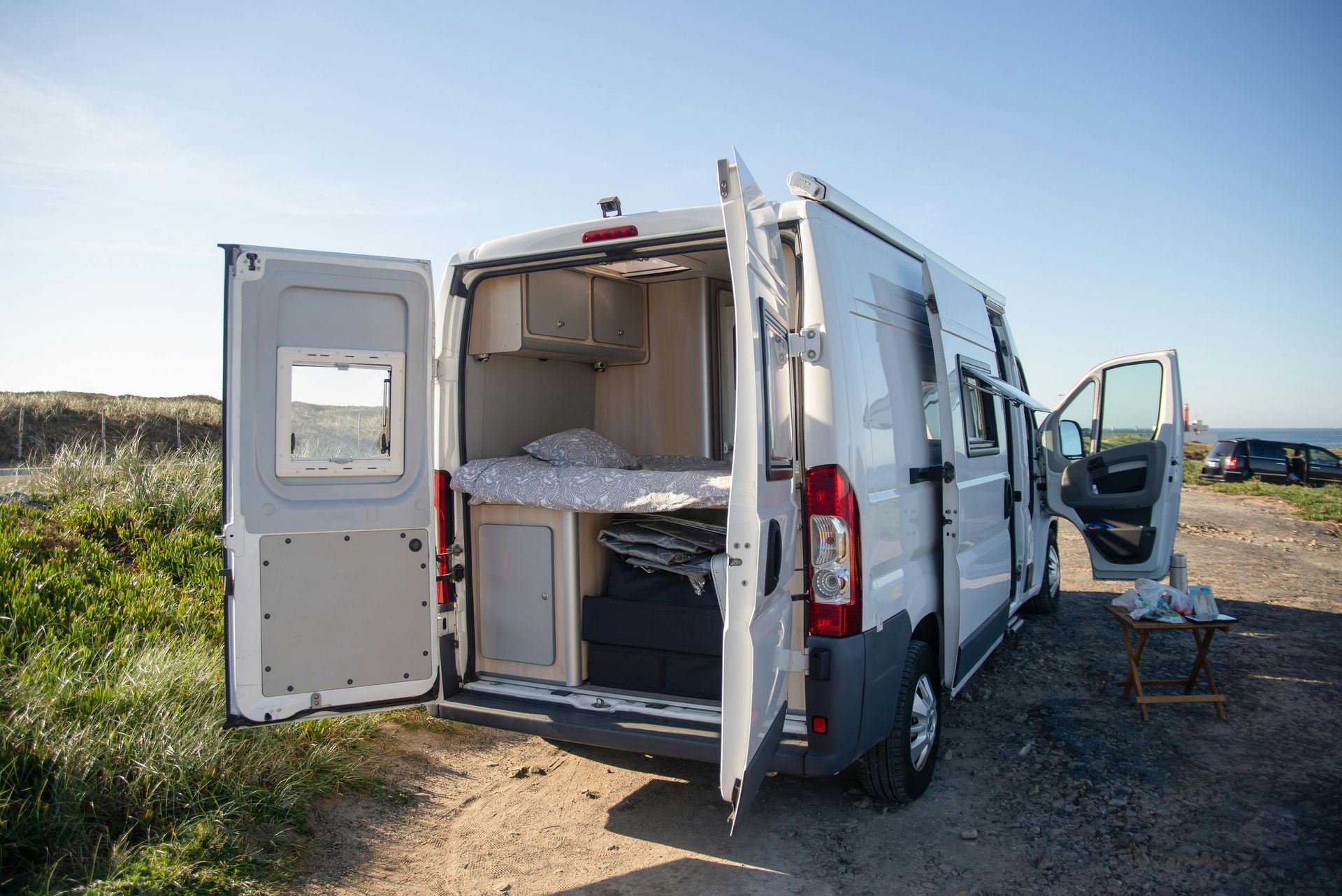 White camper van with open back doors revealing a bed, parked near grass and ocean.