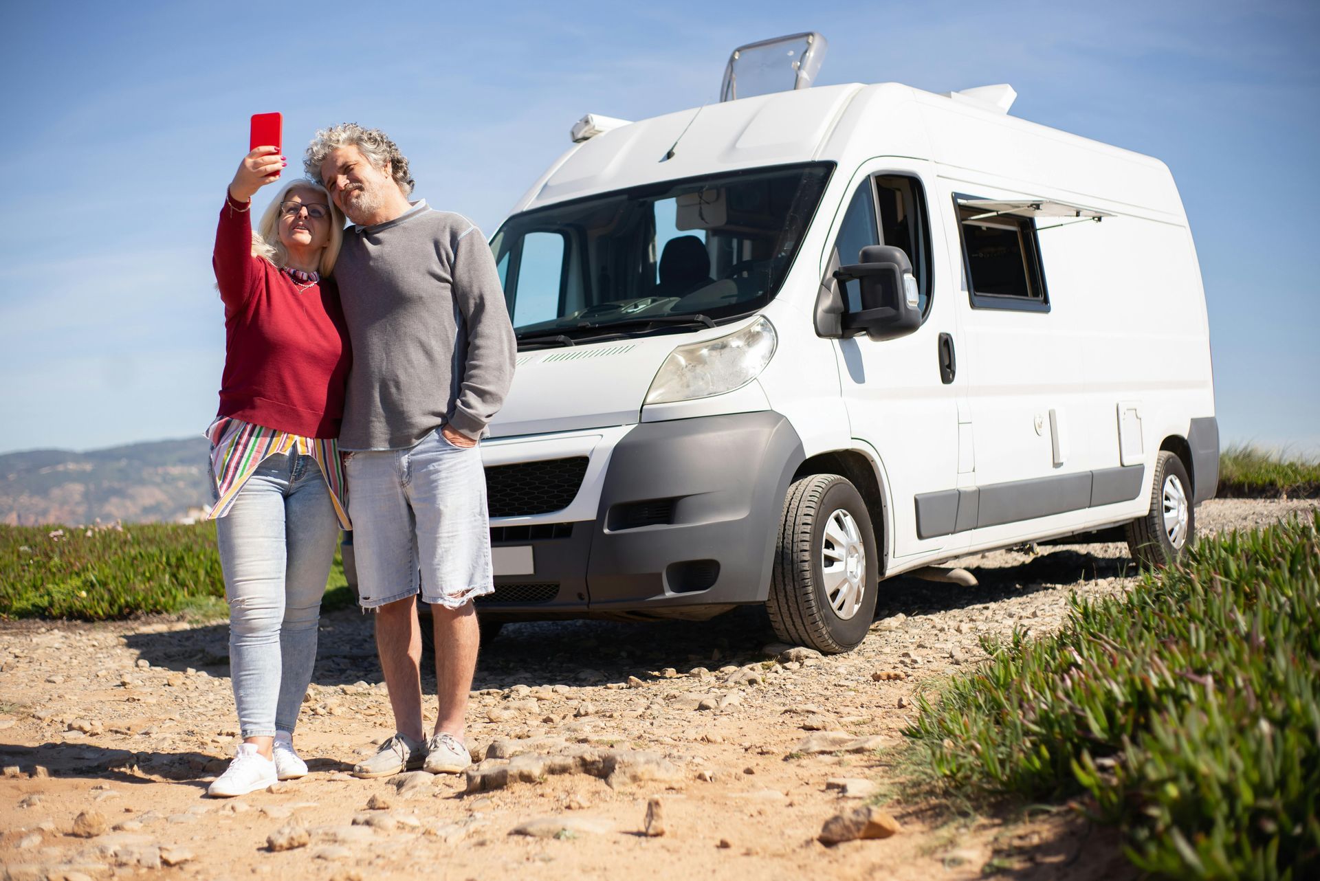 Couple taking selfie in front of a white camper van on a sunny day.