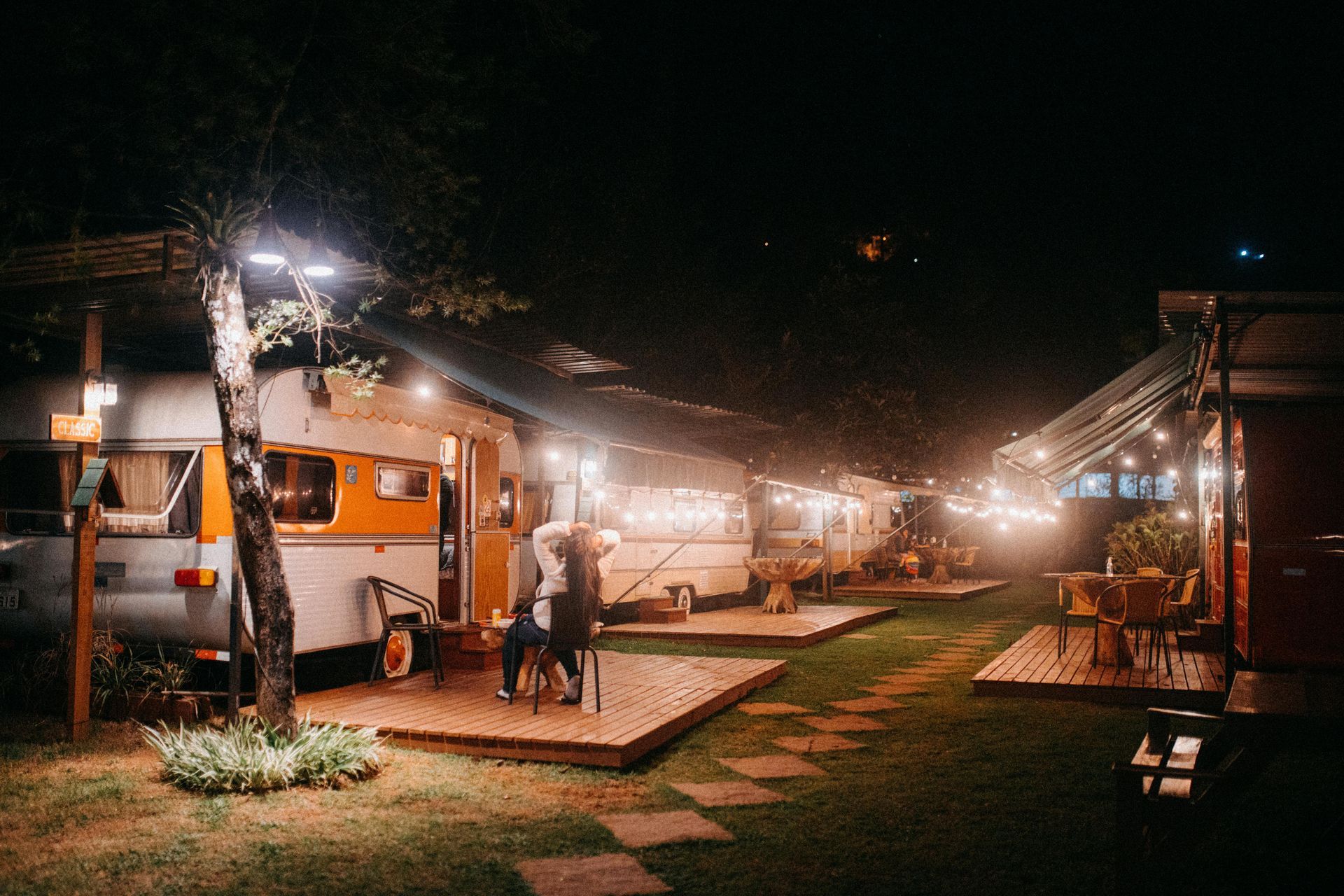 Campground at night with lit retro campers. String lights illuminate wooden decks and pathways.