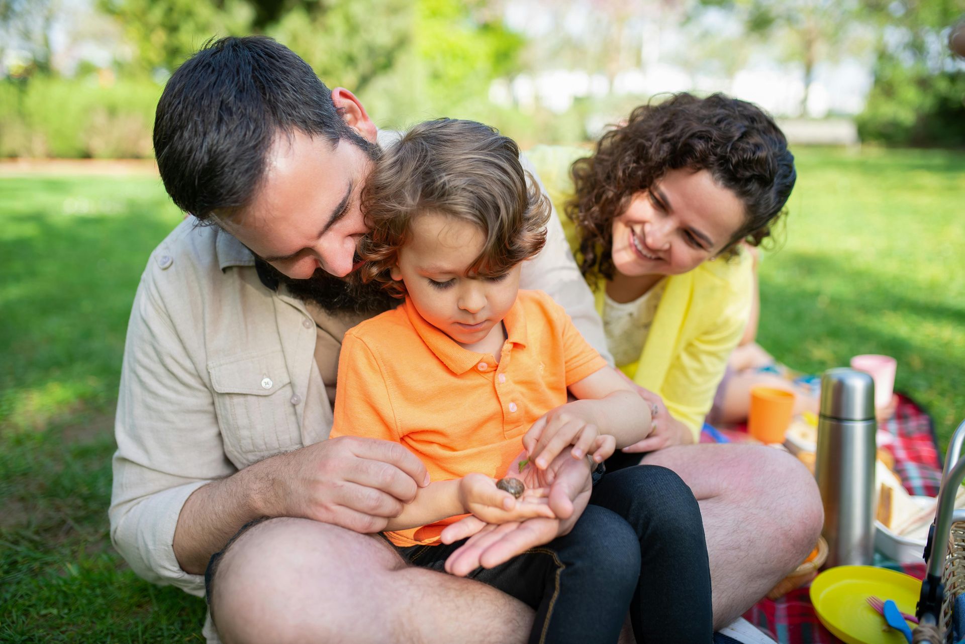 Family picnicking on a blanket in the grass. A child examines something in their hands, while the parents watch.