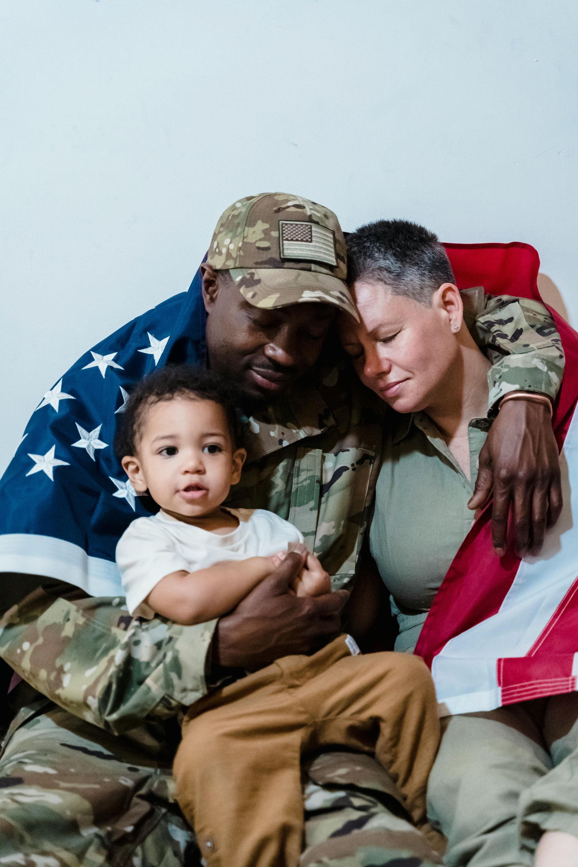 Soldier embraces family, draped in an American flag; child looks at the viewer with arms crossed.