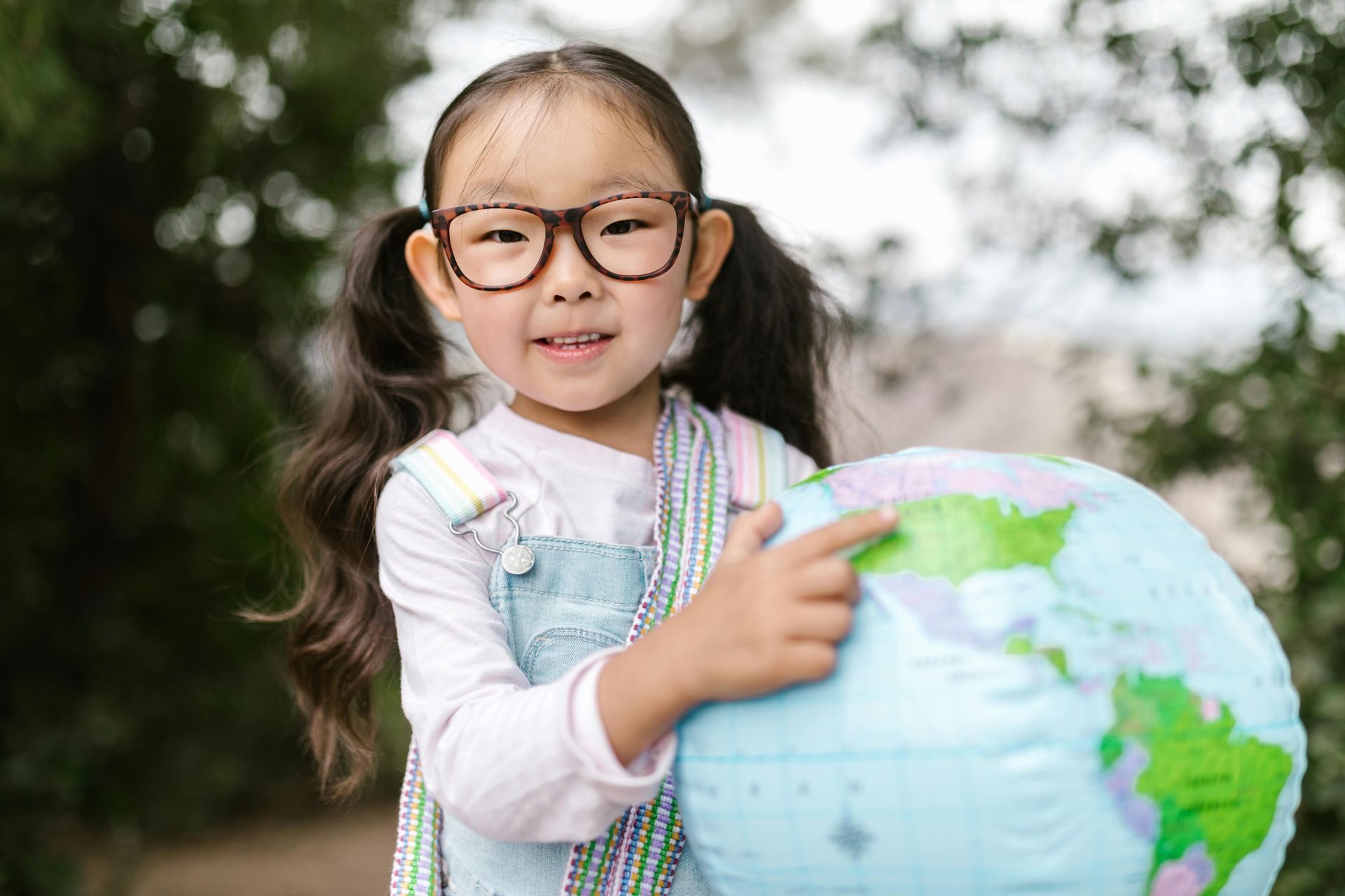 Girl with glasses pointing at a globe, smiling outside.