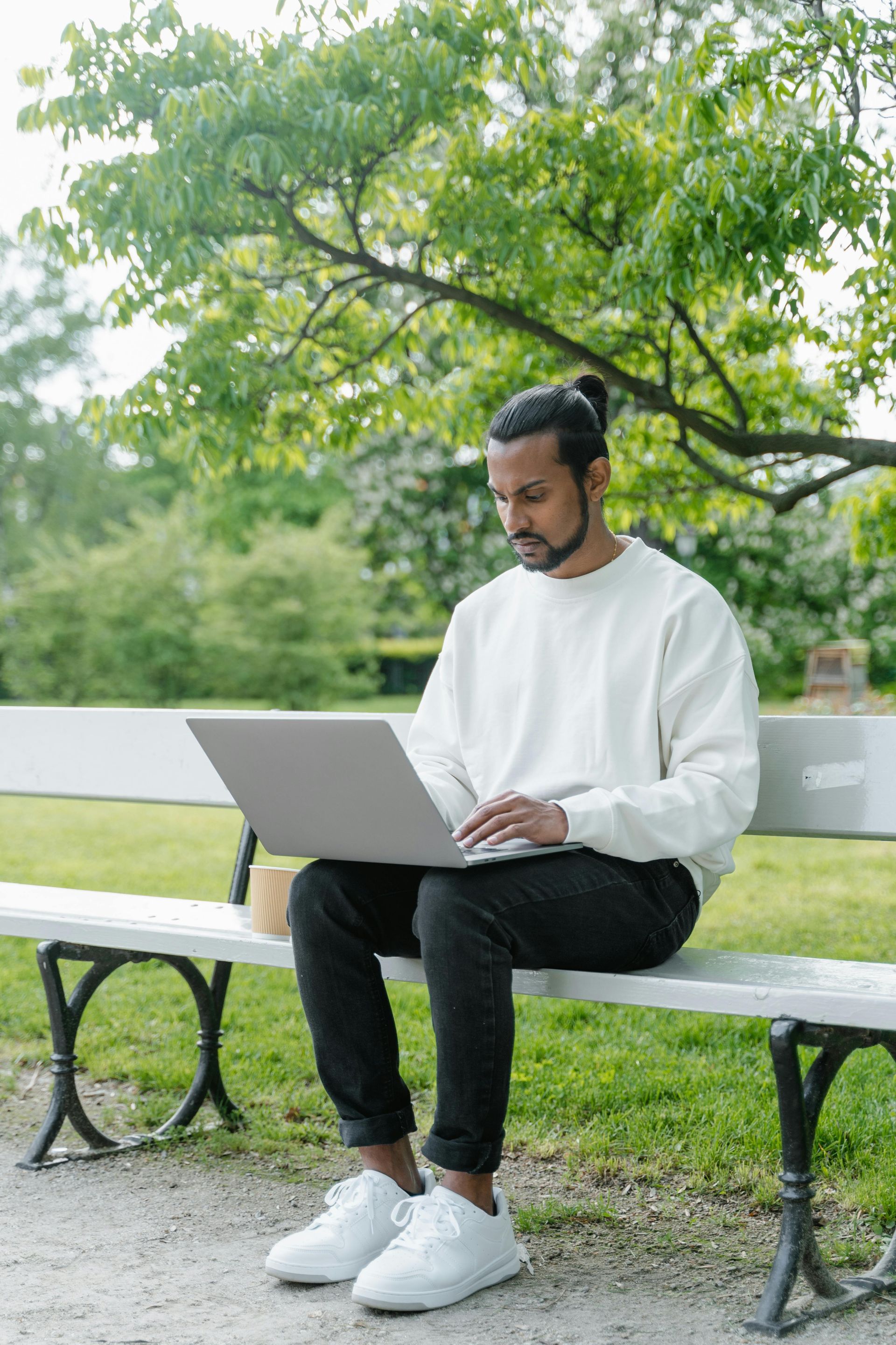Man working on laptop while sitting on a park bench under a tree.