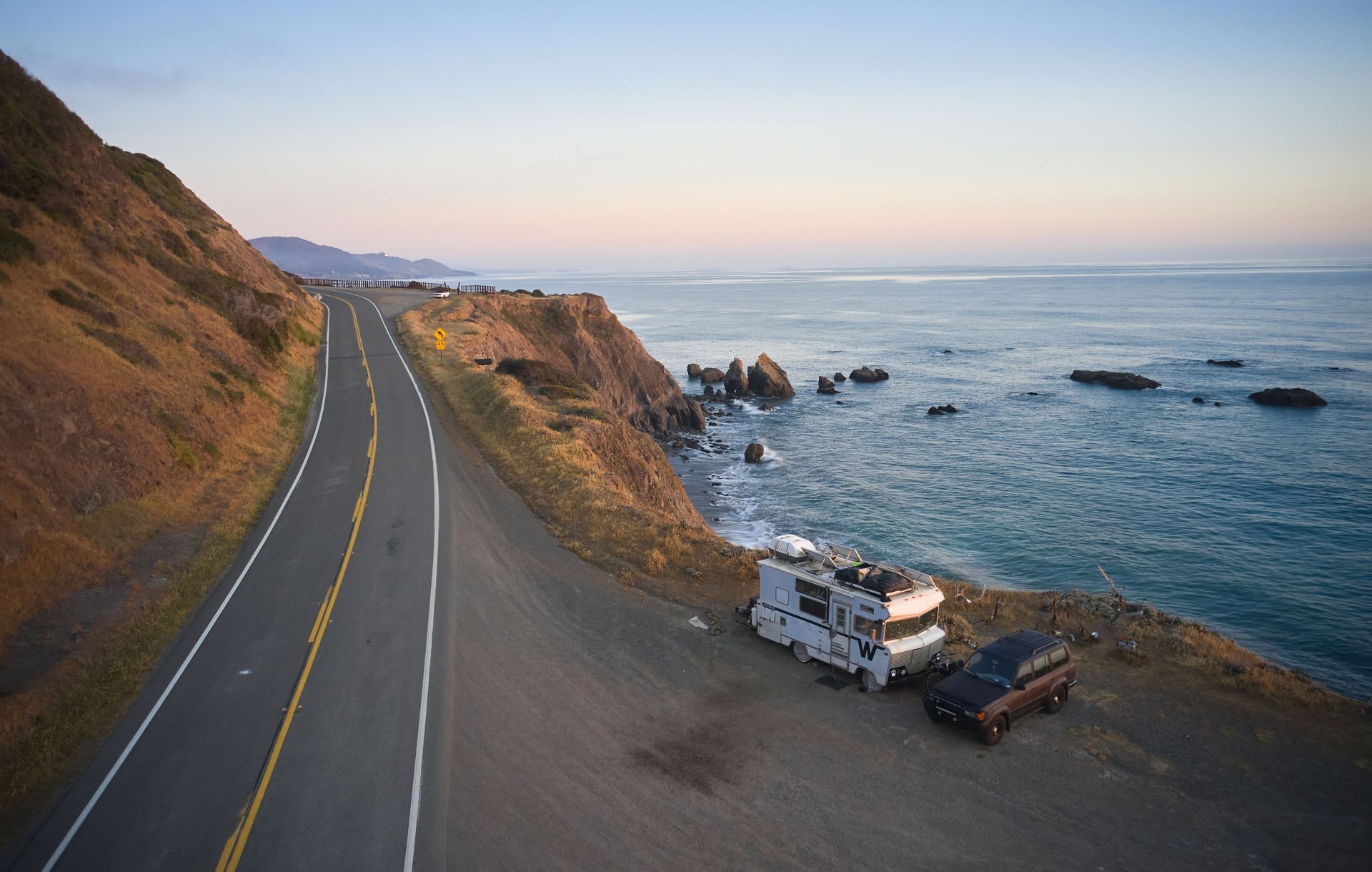 Coastal road curving along cliffs beside the ocean at sunset, with a parked RV and trailer below.