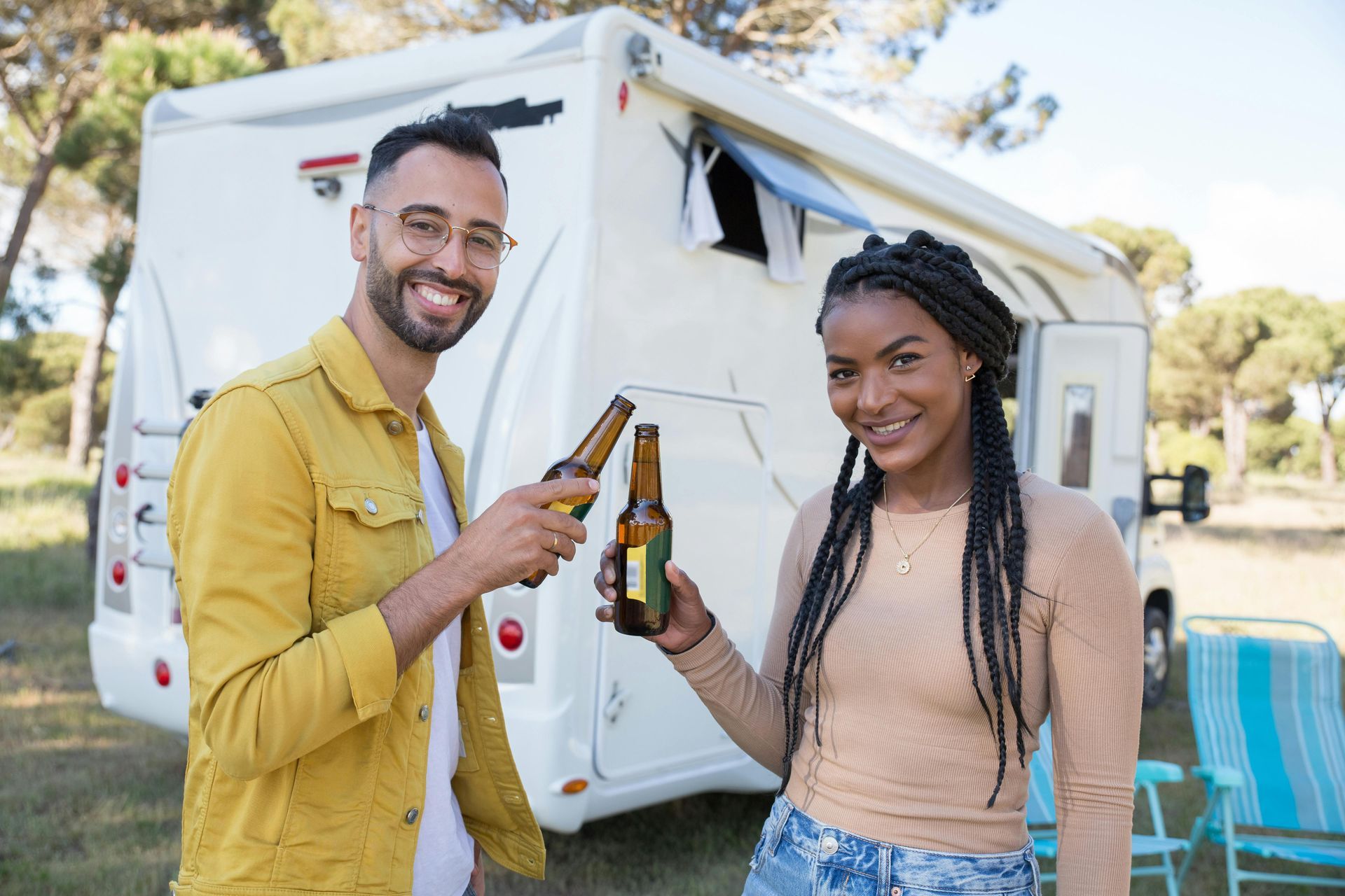 Couple toasting beer bottles, smiling in front of a campervan in a sunny outdoor setting.