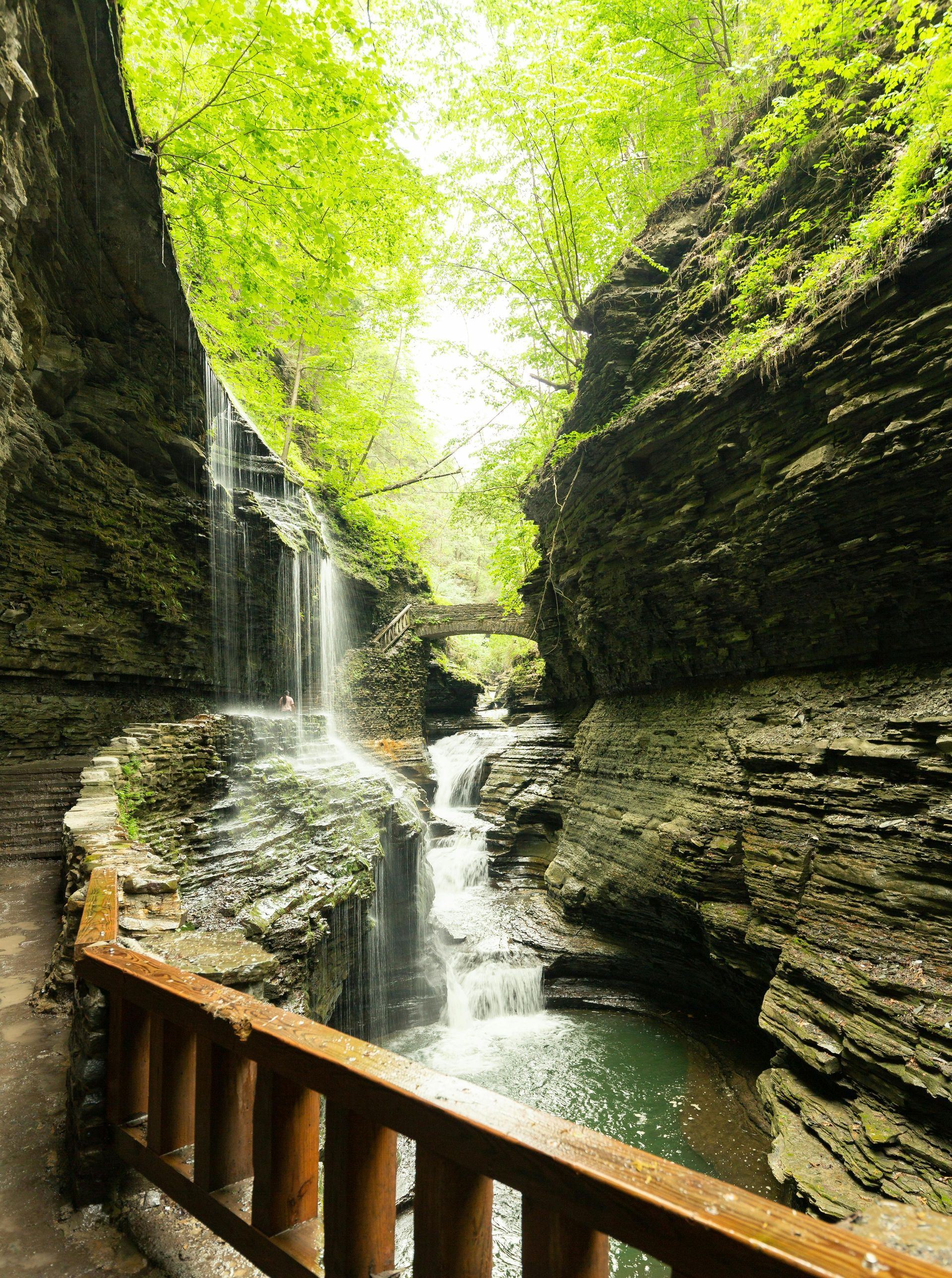 Waterfall cascading down layered rock cliffs, wooden railing in foreground, lush green foliage.