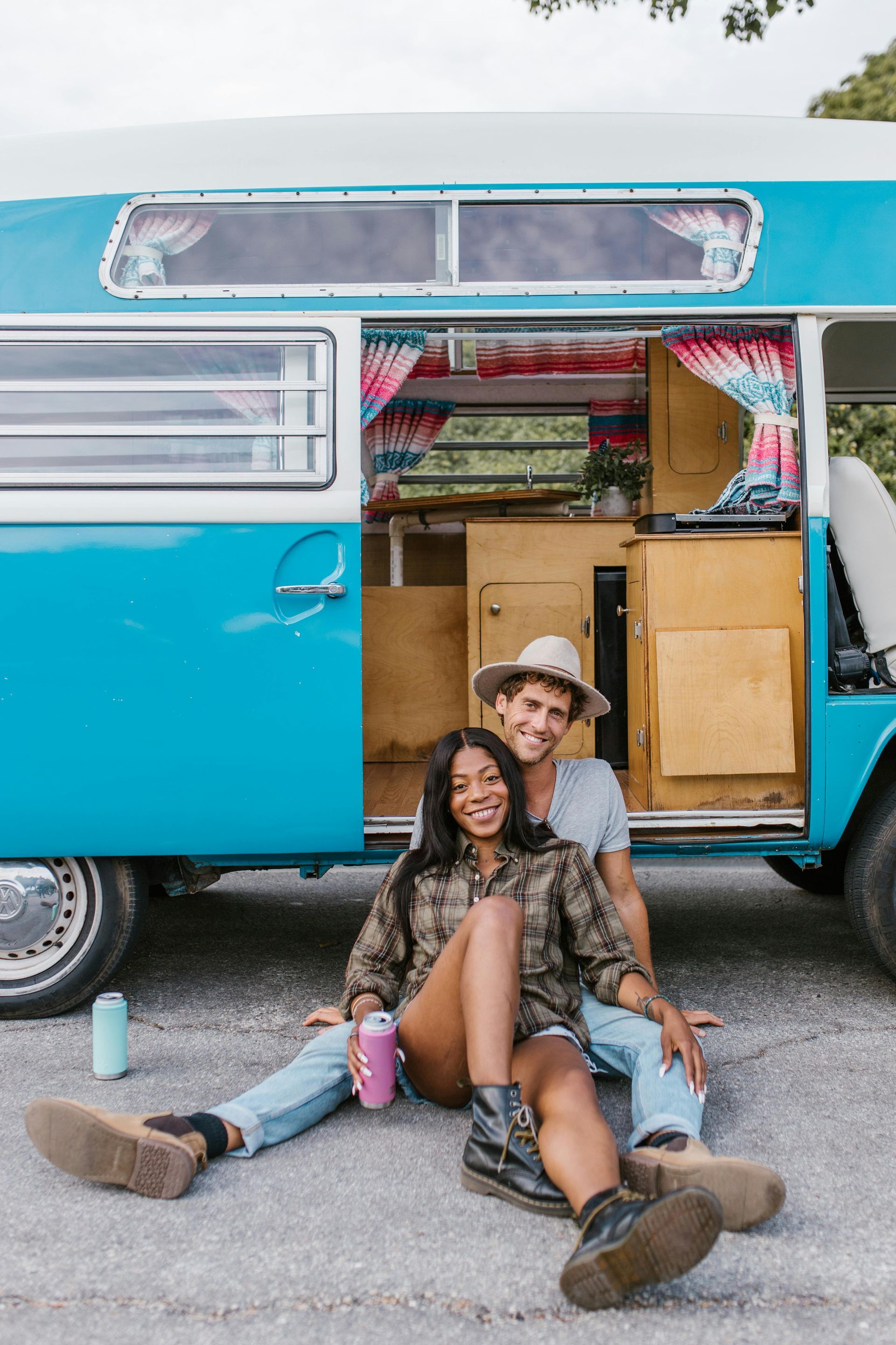 Couple sits in front of blue campervan. One wears a hat. Both are smiling.