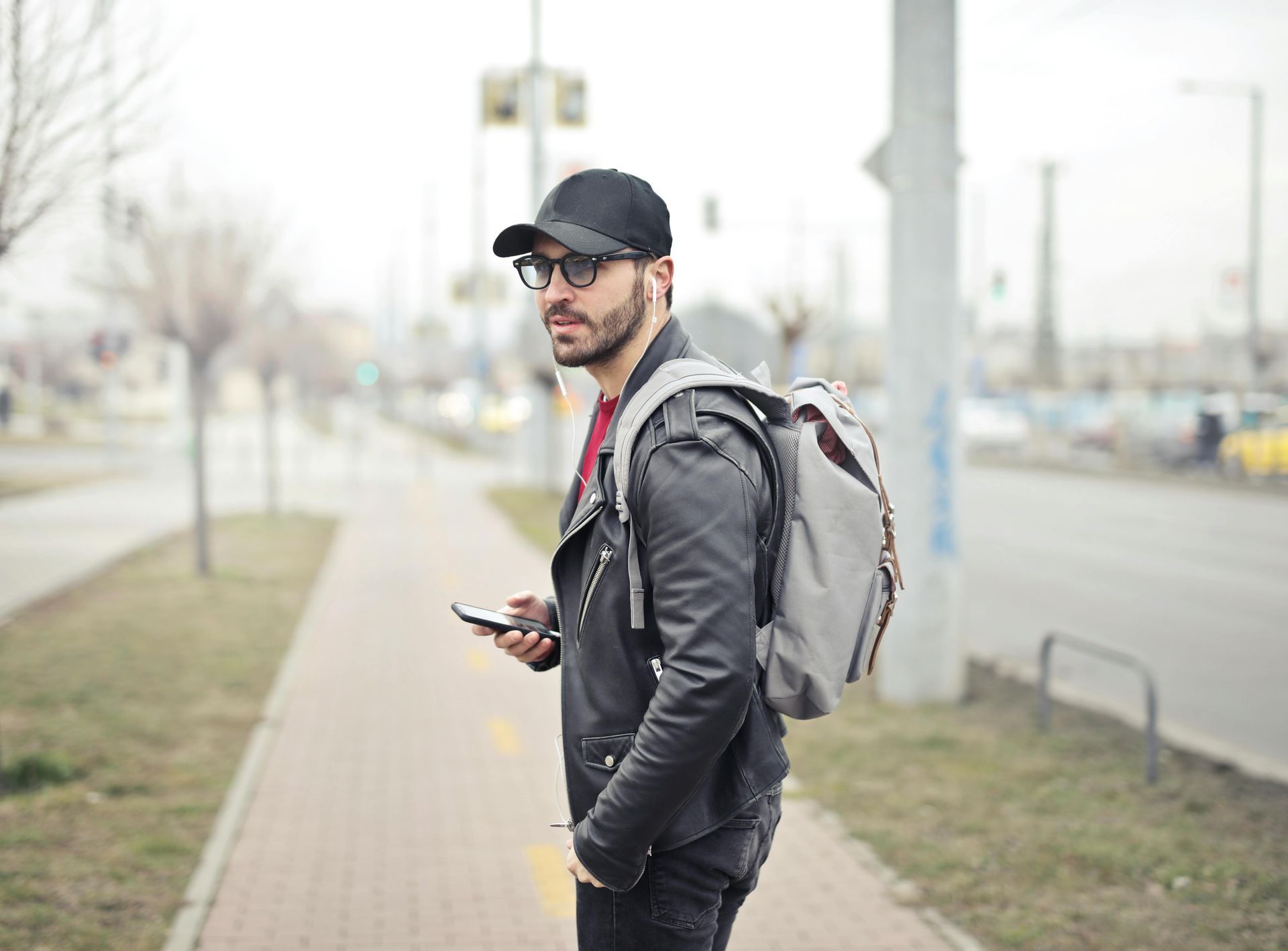 Man in black jacket, hat, and backpack looks over shoulder, holding phone, standing on sidewalk.