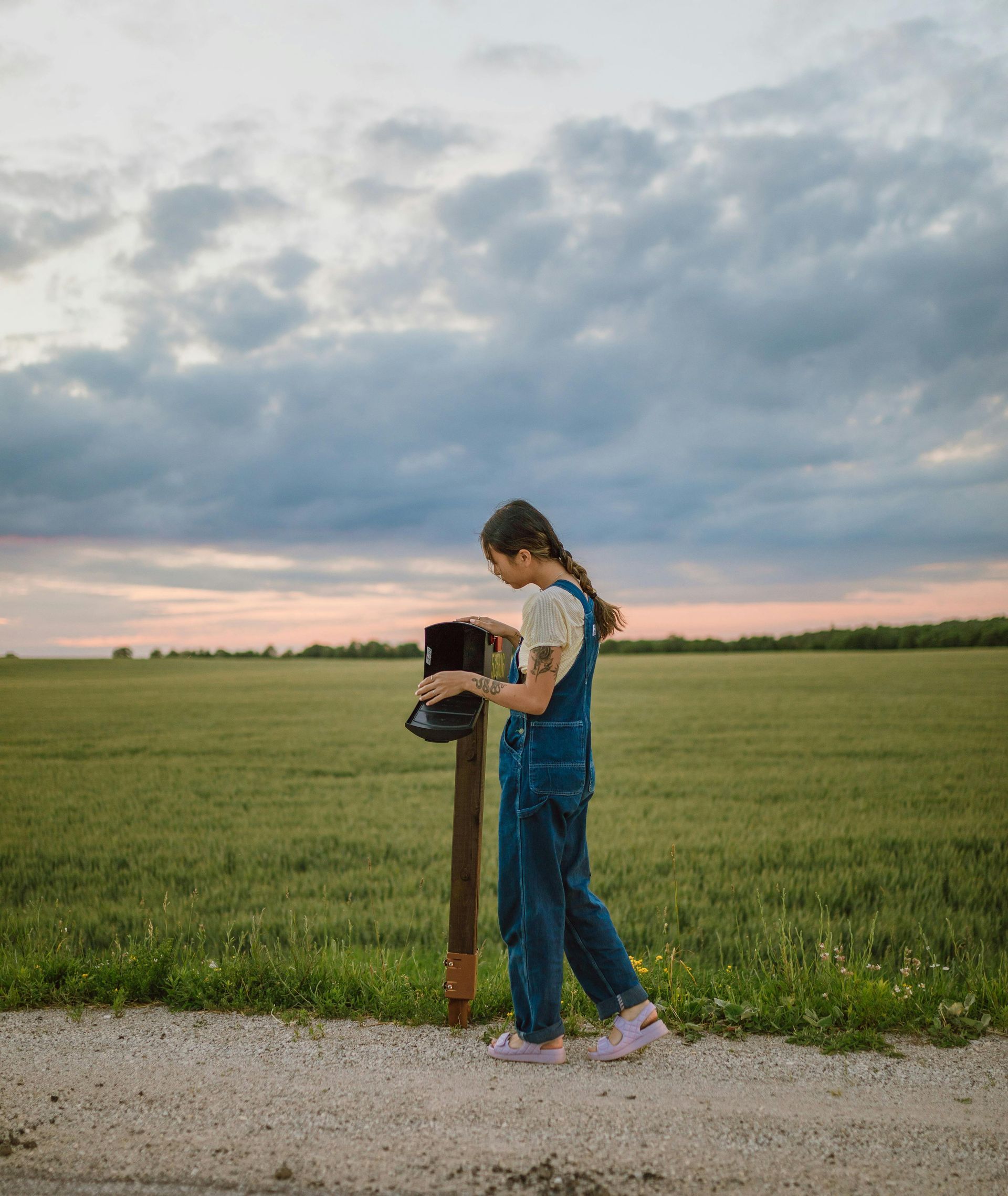 Person in denim overalls at a rural mailbox, grassy field backdrop, cloudy sky.