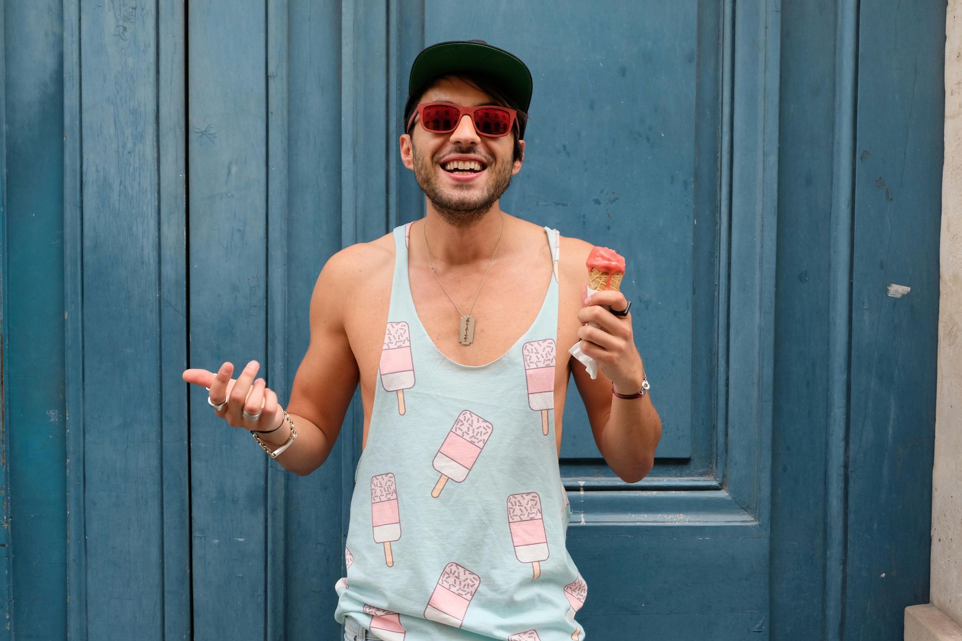 Man in a tank top with popsicle pattern, holding ice cream, and smiling in front of a blue door.