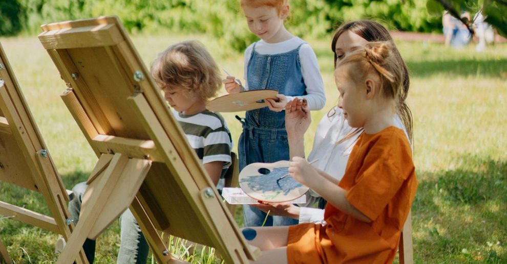 Children and a teacher painting at easels outdoors in a sunny park.