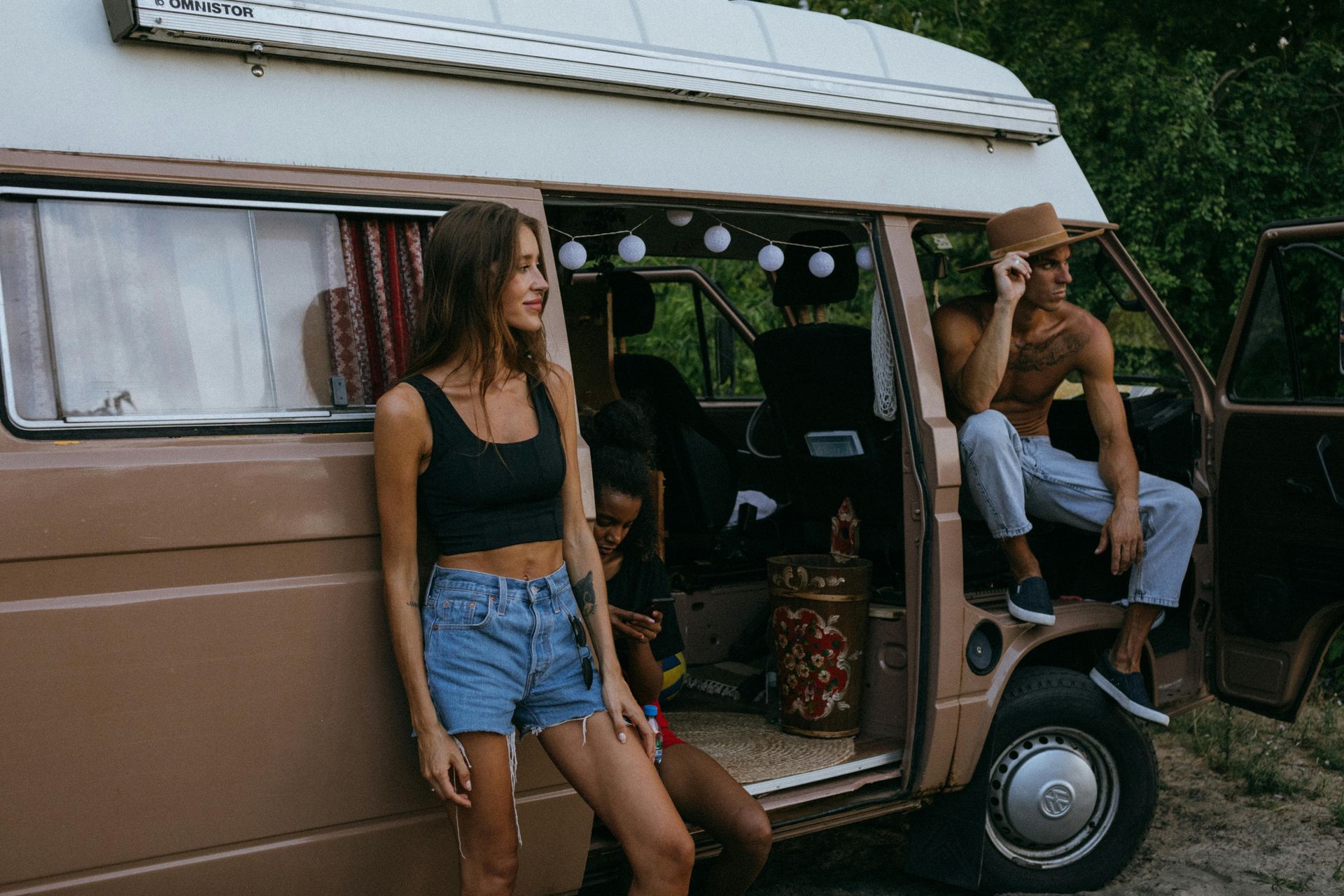 People relaxing inside a brown van. A woman and a man sit outside, while another person is inside the van.