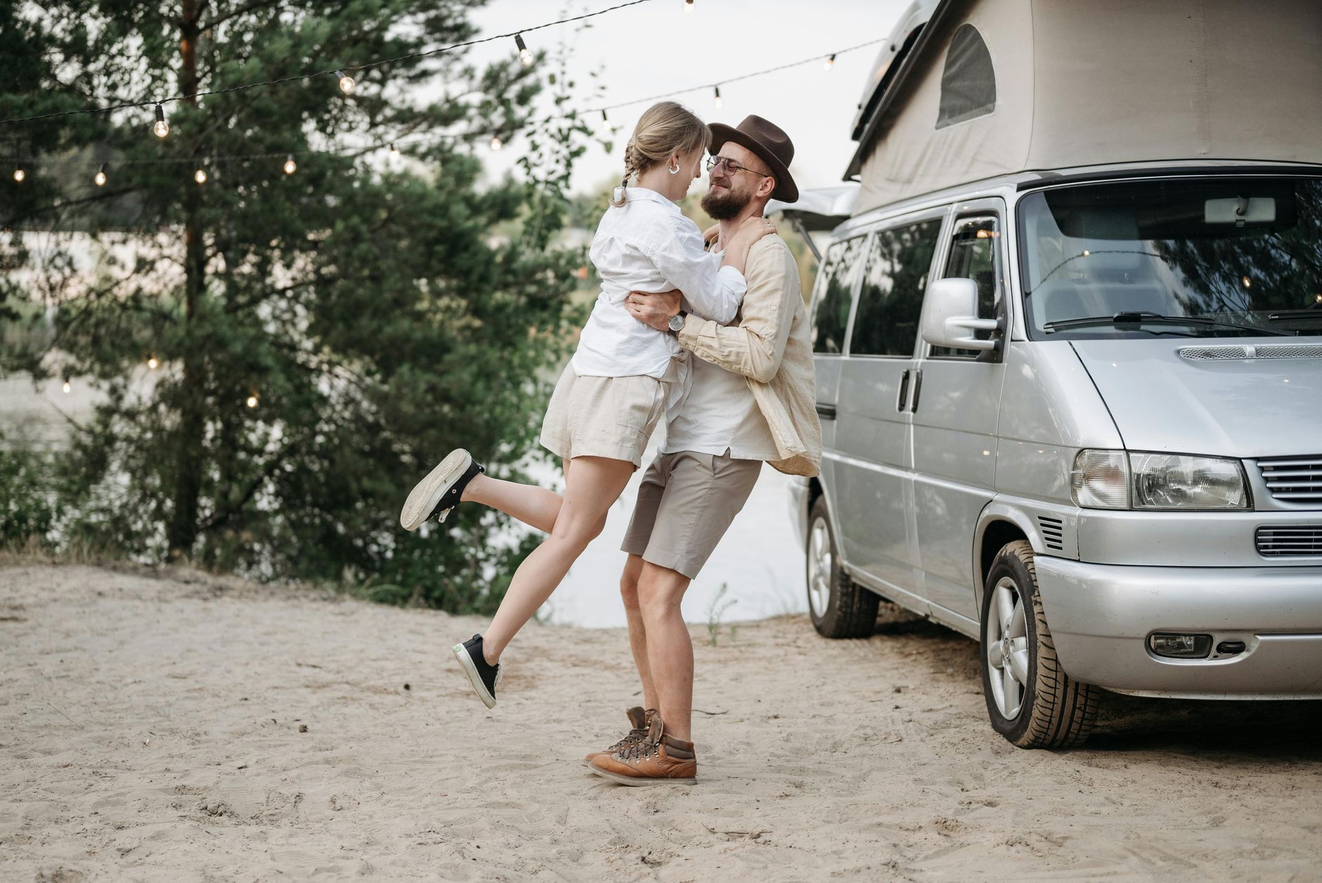 Couple embraces near a camper van. Woman lifted in the air by man. Outdoors with trees and string lights.