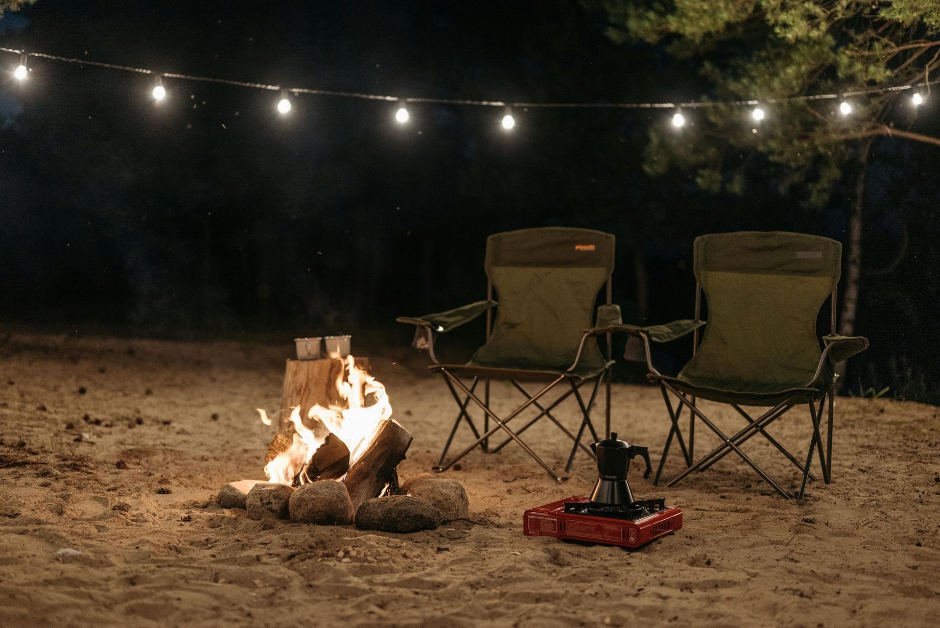 Two camping chairs by a campfire, string lights overhead. Coffee maker on a red stove, outdoors at night.