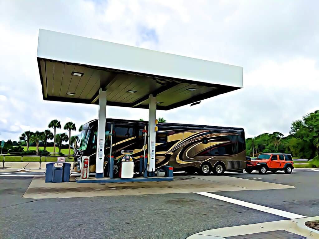 Large RV and car at gas pump under a white canopy; palm trees and cloudy sky.
