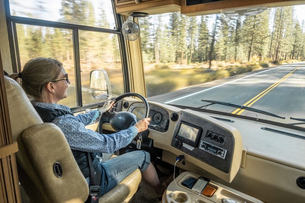 Woman driving an RV on a road lined with trees. She smiles and wears sunglasses.