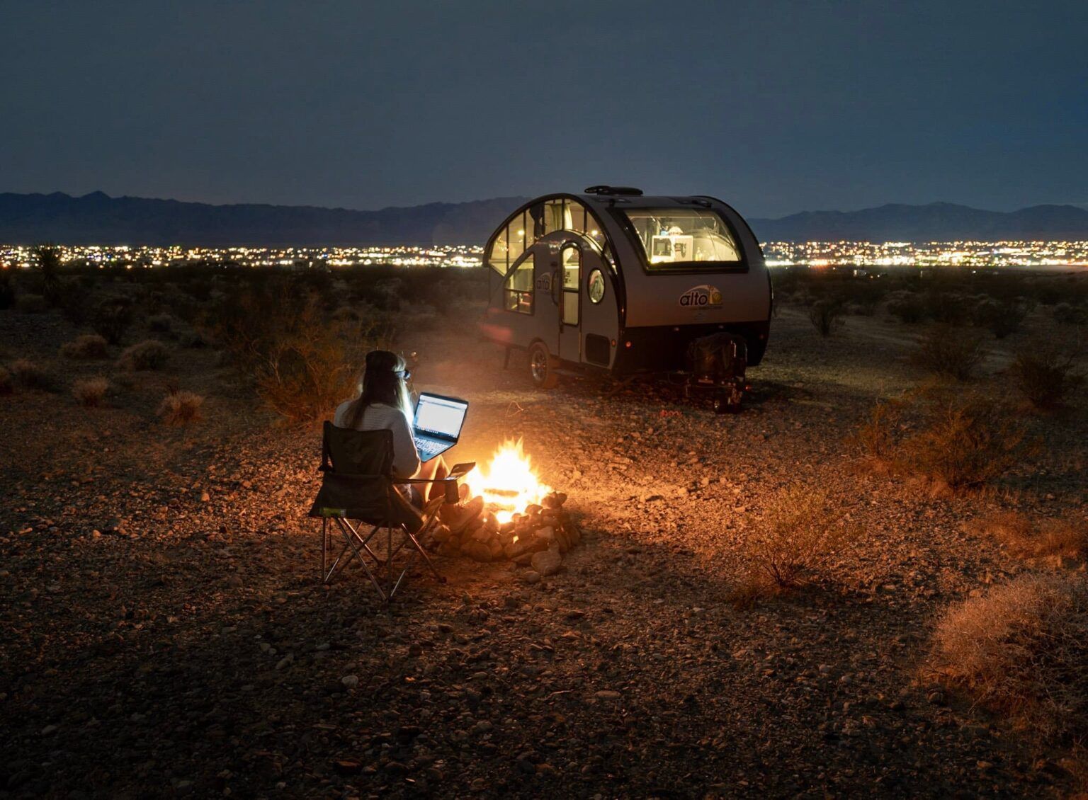 Woman working on laptop by campfire near teardrop trailer at night, city lights in the distance.
