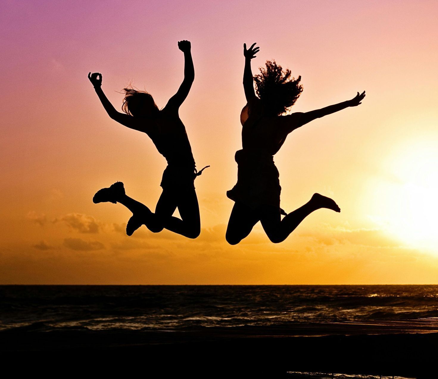 Silhouetted figures of two people jumping for joy at sunset over the ocean.