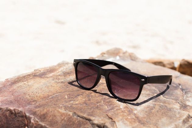 Occhiali da sole neri poggiano su una roccia marrone con uno sfondo sfocato di spiaggia sabbiosa.