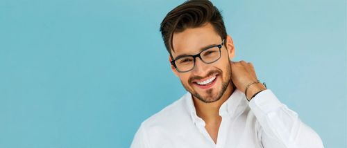 Un uomo con gli occhiali sorride, si tocca il collo e indossa una camicia bianca, su uno sfondo azzurro.
