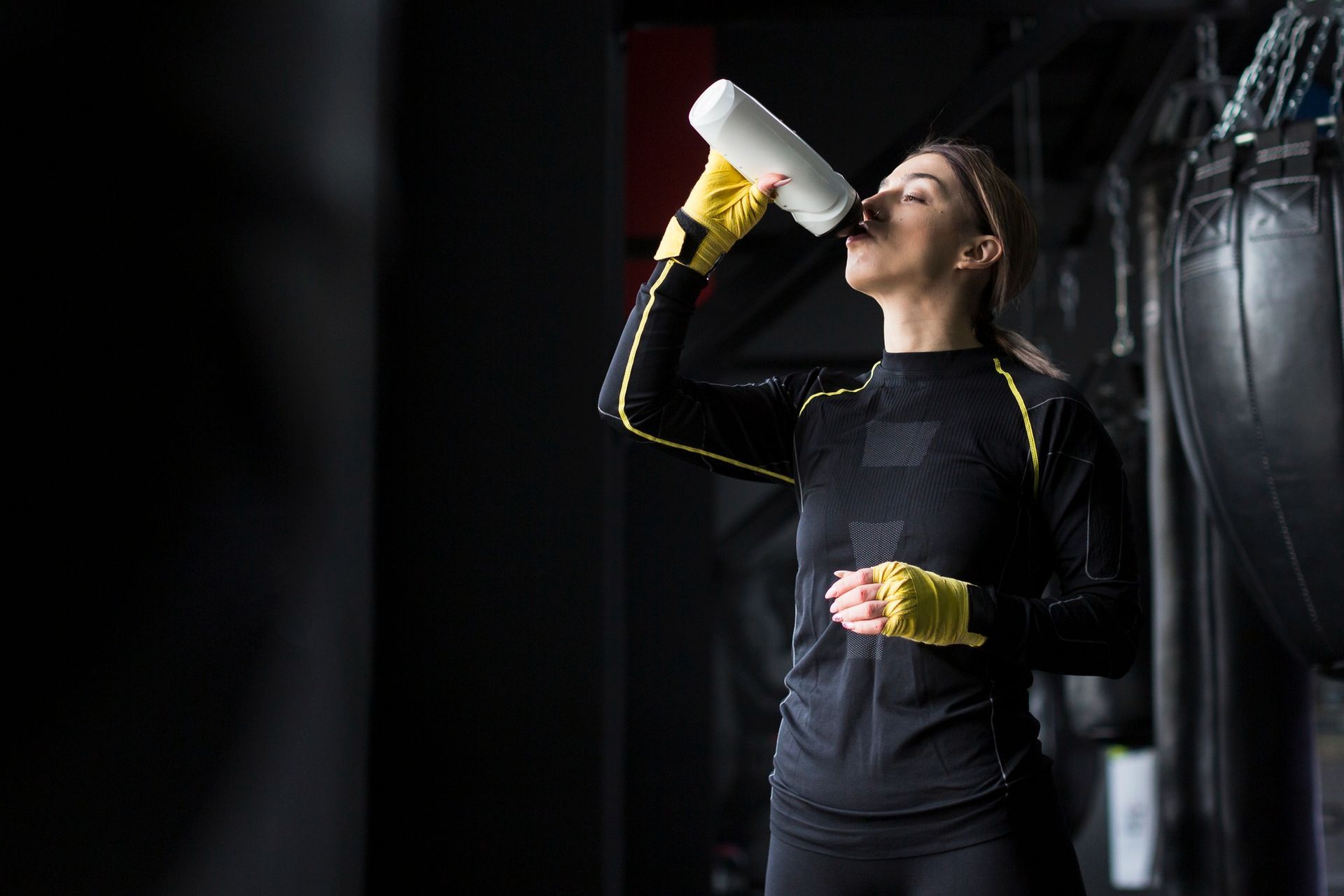 Una mujer bebe de una botella de agua blanca en un gimnasio oscuro. Lleva ropa deportiva negra con detalles amarillos.
