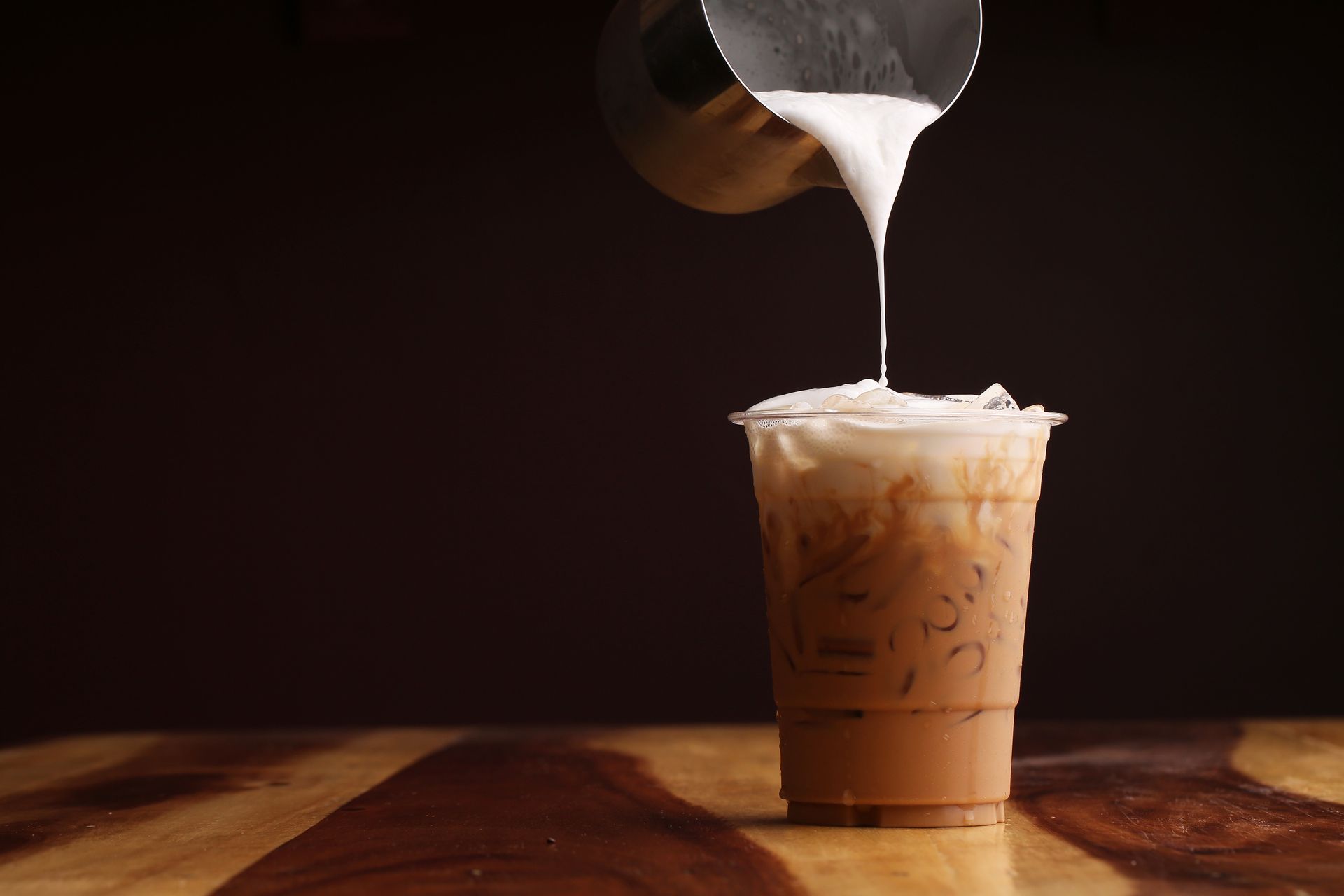 Iced coffee is being poured into a plastic cup on a wooden table.