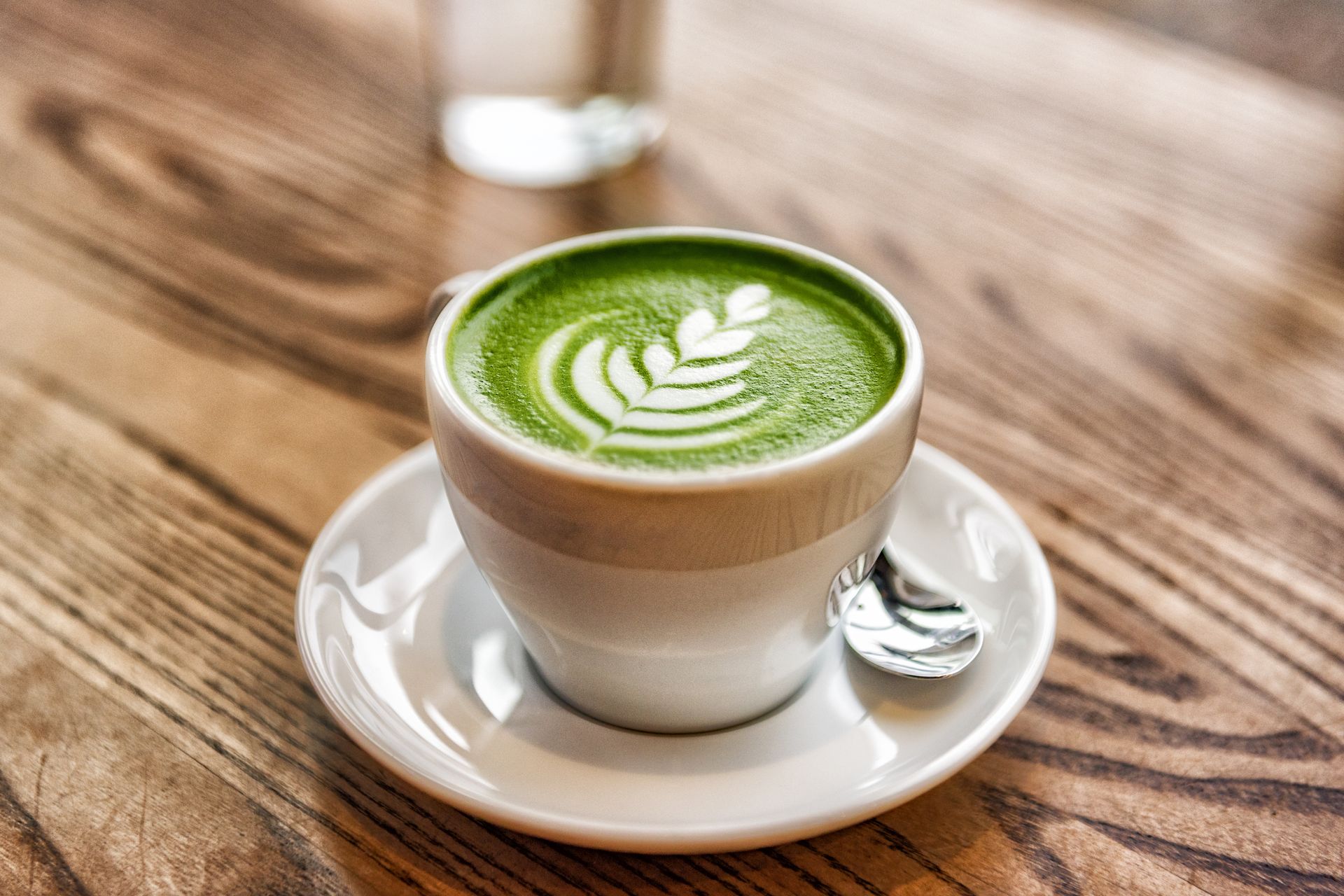 A cup of green tea latte on a saucer with a spoon on a wooden table.