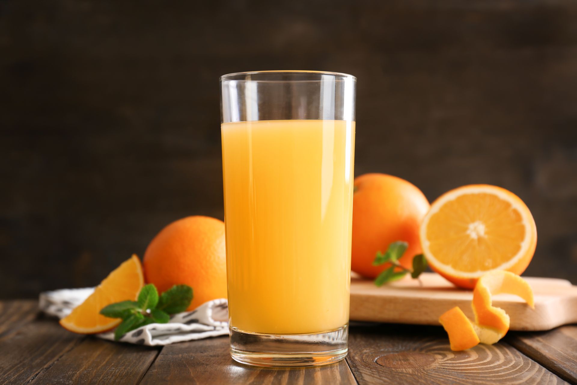 A glass of orange juice is sitting on a wooden table next to oranges.