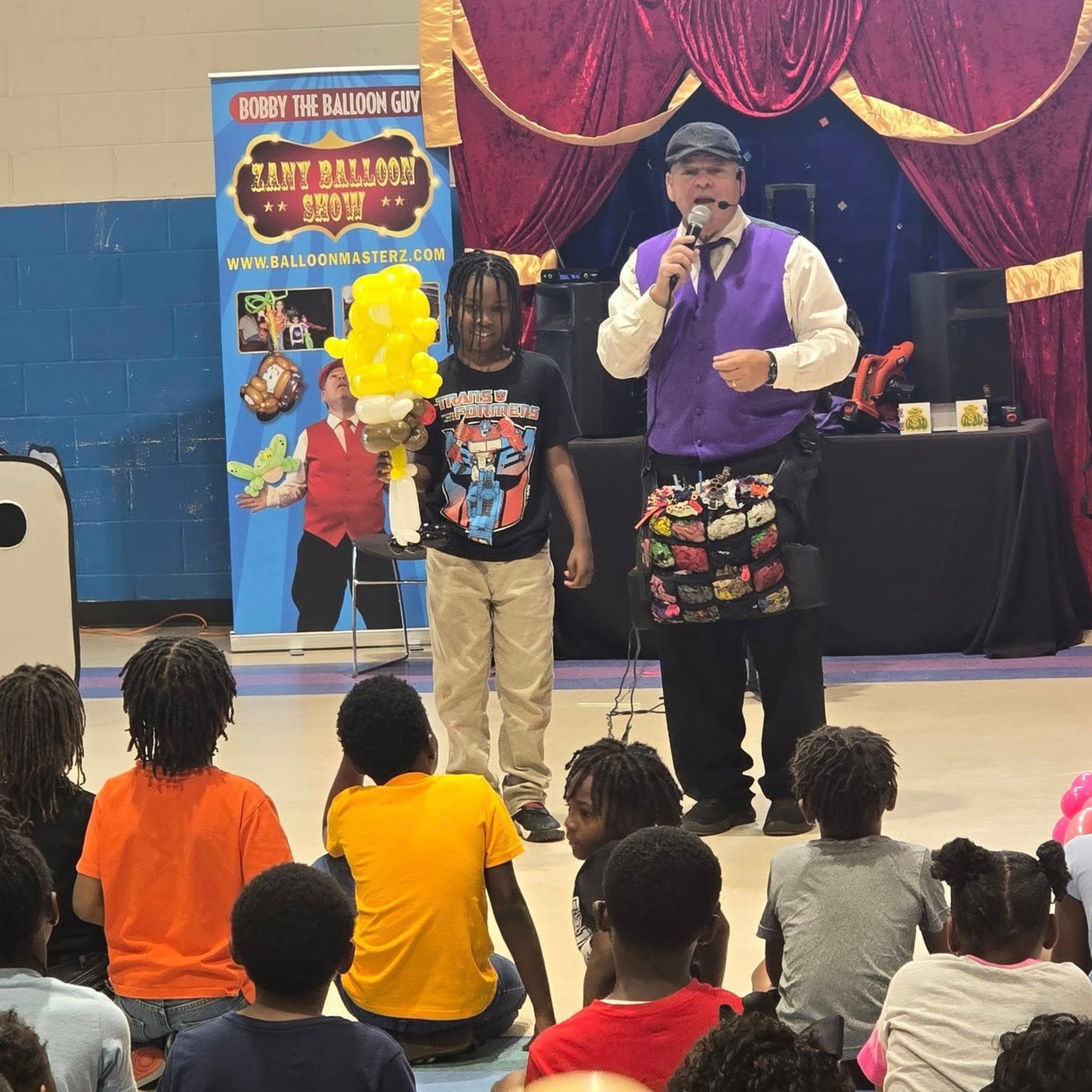 Magician and boy on stage with balloon creation, performing for children in a school gym.