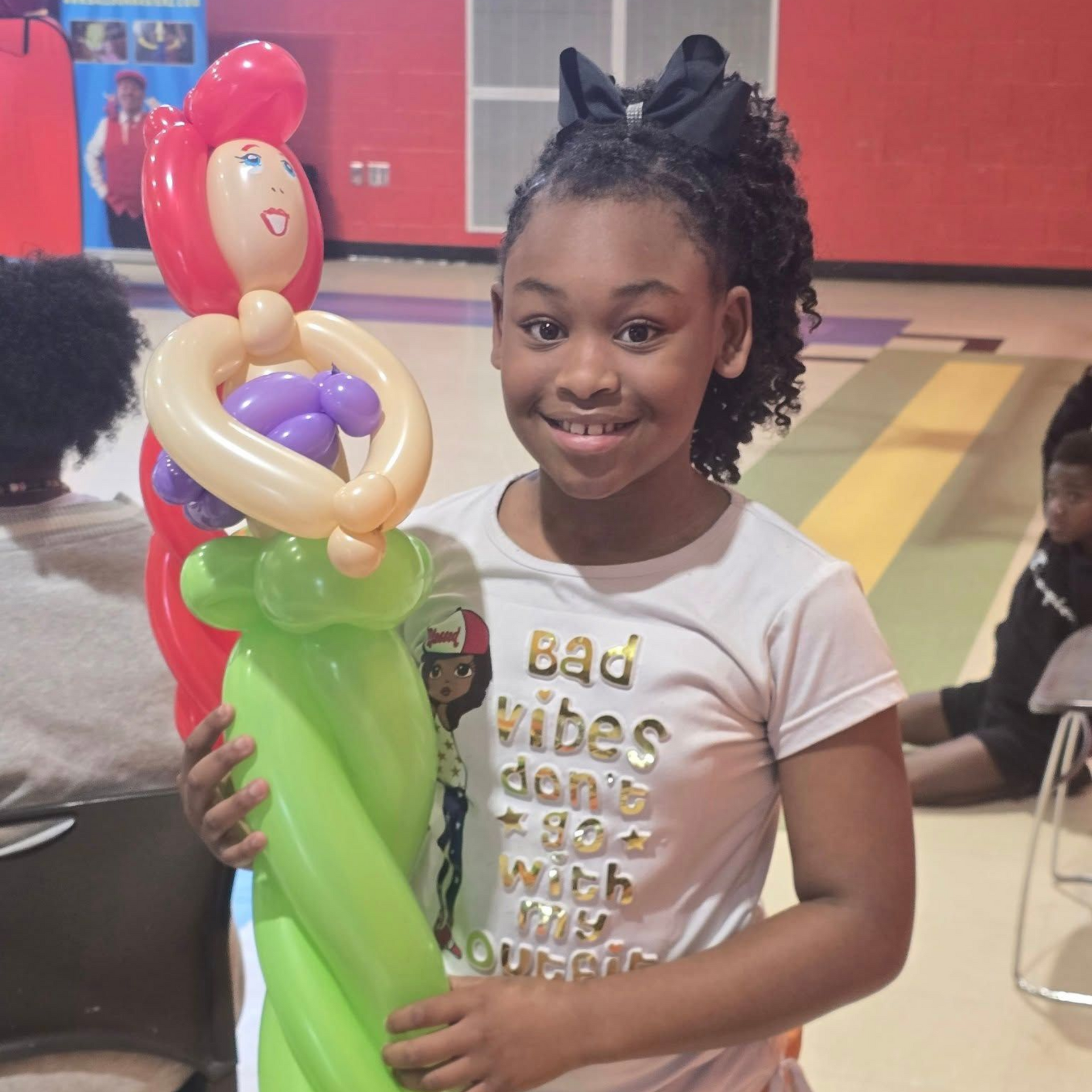 Girl holding balloon sculpture of a mermaid; wearing white shirt with black bow; smiles in indoor setting.