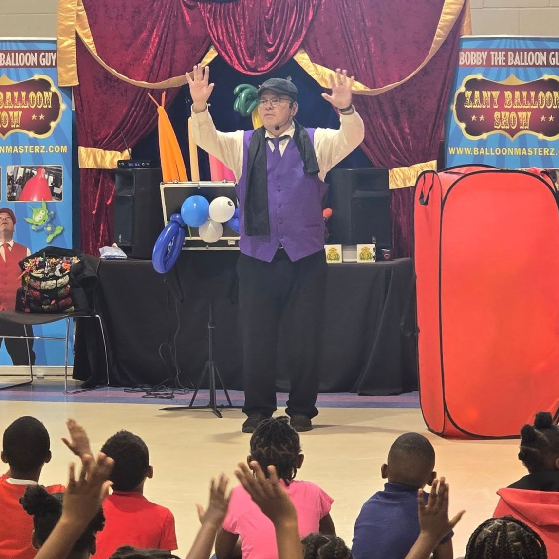Magician in purple vest performing on stage with raised hands, audience in foreground.