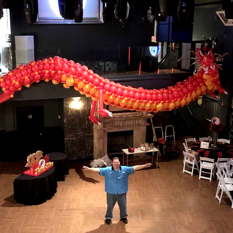 Girl holding balloon sculpture of a mermaid; wearing white shirt with black bow; smiles in indoor setting.