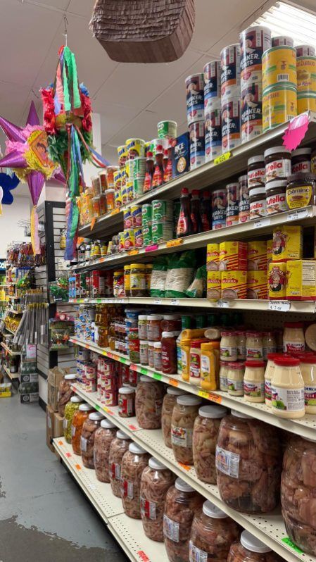 A grocery aisle with shelves of packaged foods, jars, and colorful piñatas hanging on the left.