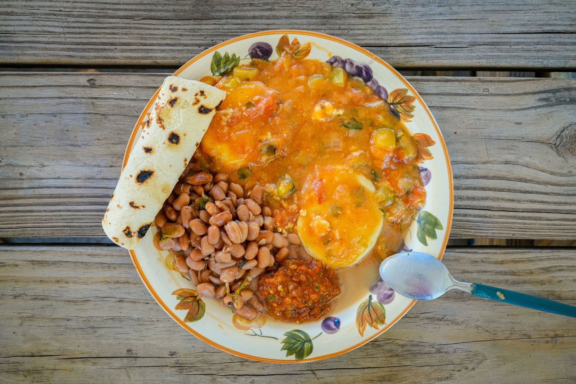Plate of saucy food with beans and bread on a rustic wooden table, with a spoon nearby