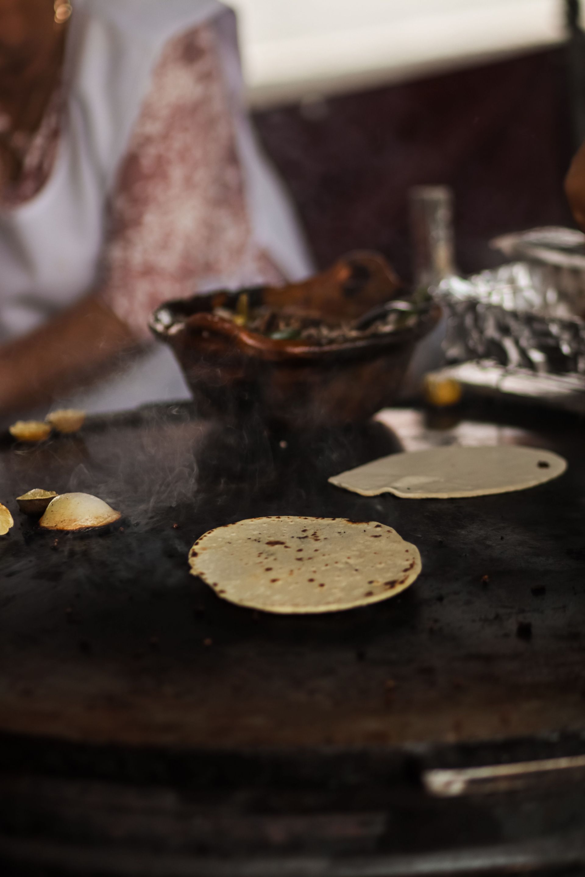 Flatbread cooking on a hot griddle with a bowl of batter and steam rising nearby