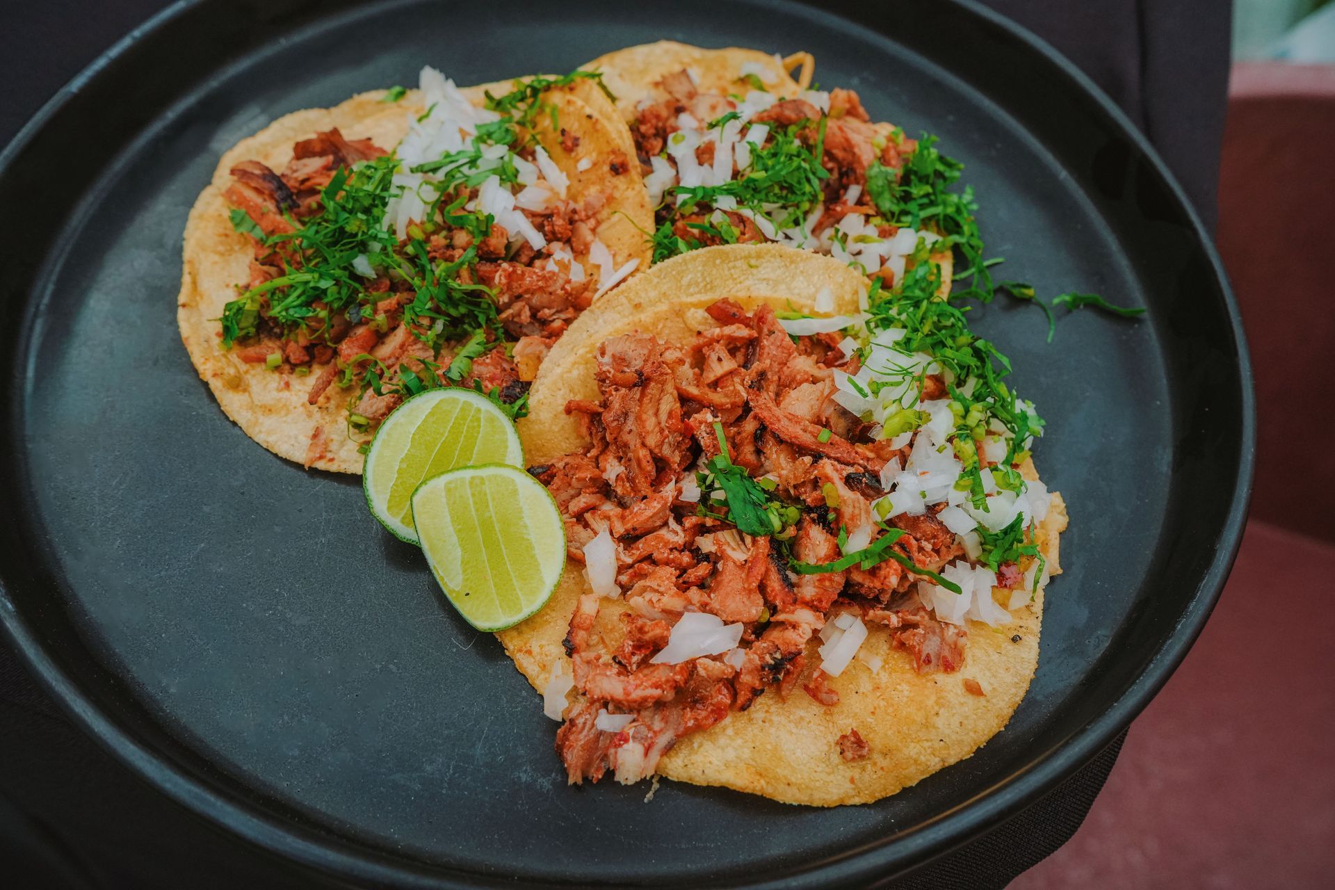 Three tacos on a black plate with lime wedges and chopped herbs