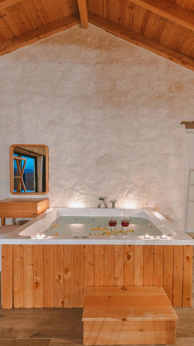 Wooden-paneled bathtub filled with water, candles, and flower petals in a rustic white-walled room with a wooden ceiling.