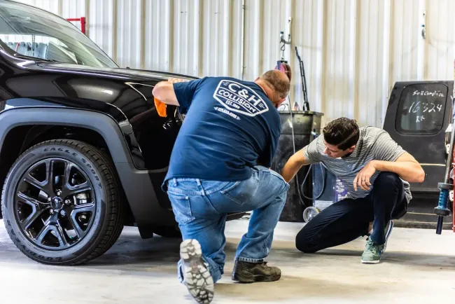 Two people working on a black car in a garage. One sprays the car while the other examines it.