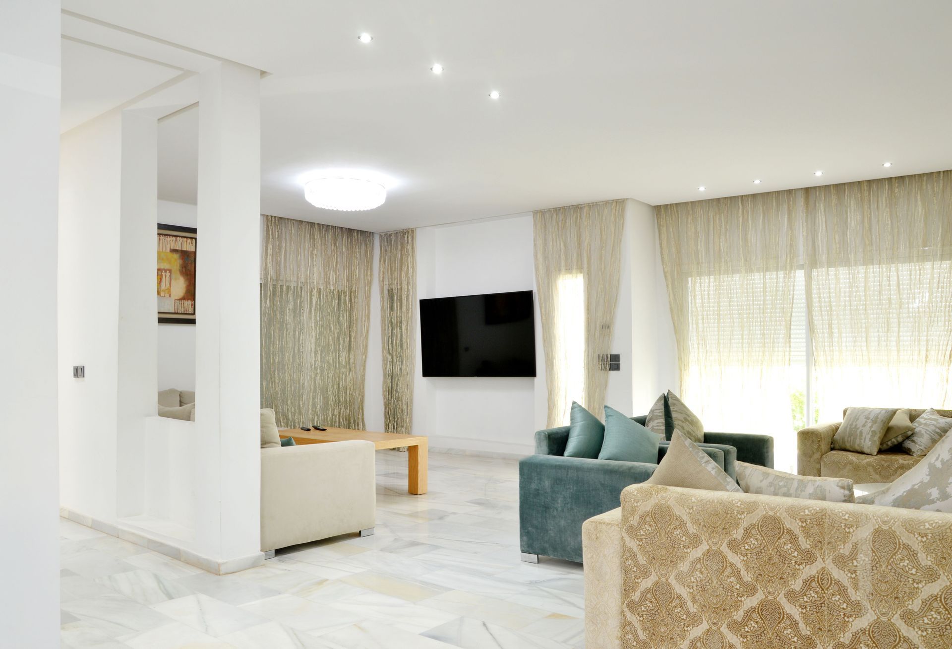 Bright modern living room with teal sofas, beige curtains, wall-mounted TV, and white marble floor